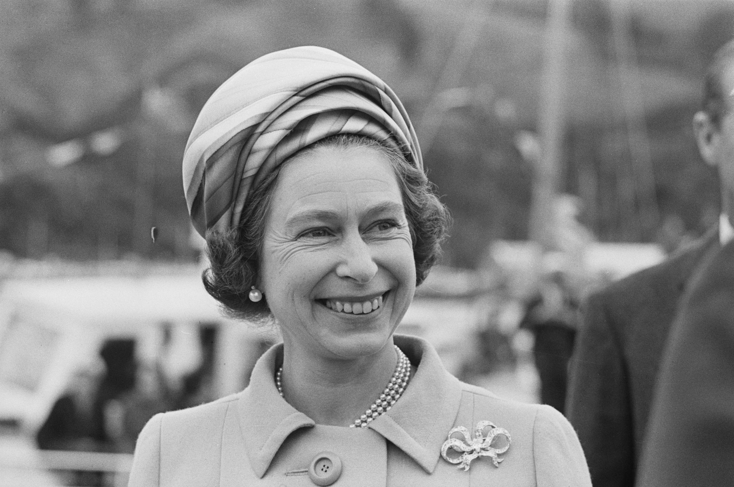 Captured in a candid moment during her 1970 visit to New Zealand, Queen Elizabeth II beams beneath a patterned headscarf and pearl necklace. Her tailored coat and delicate brooch complete the look as she charms the Commonwealth with her signature grace.