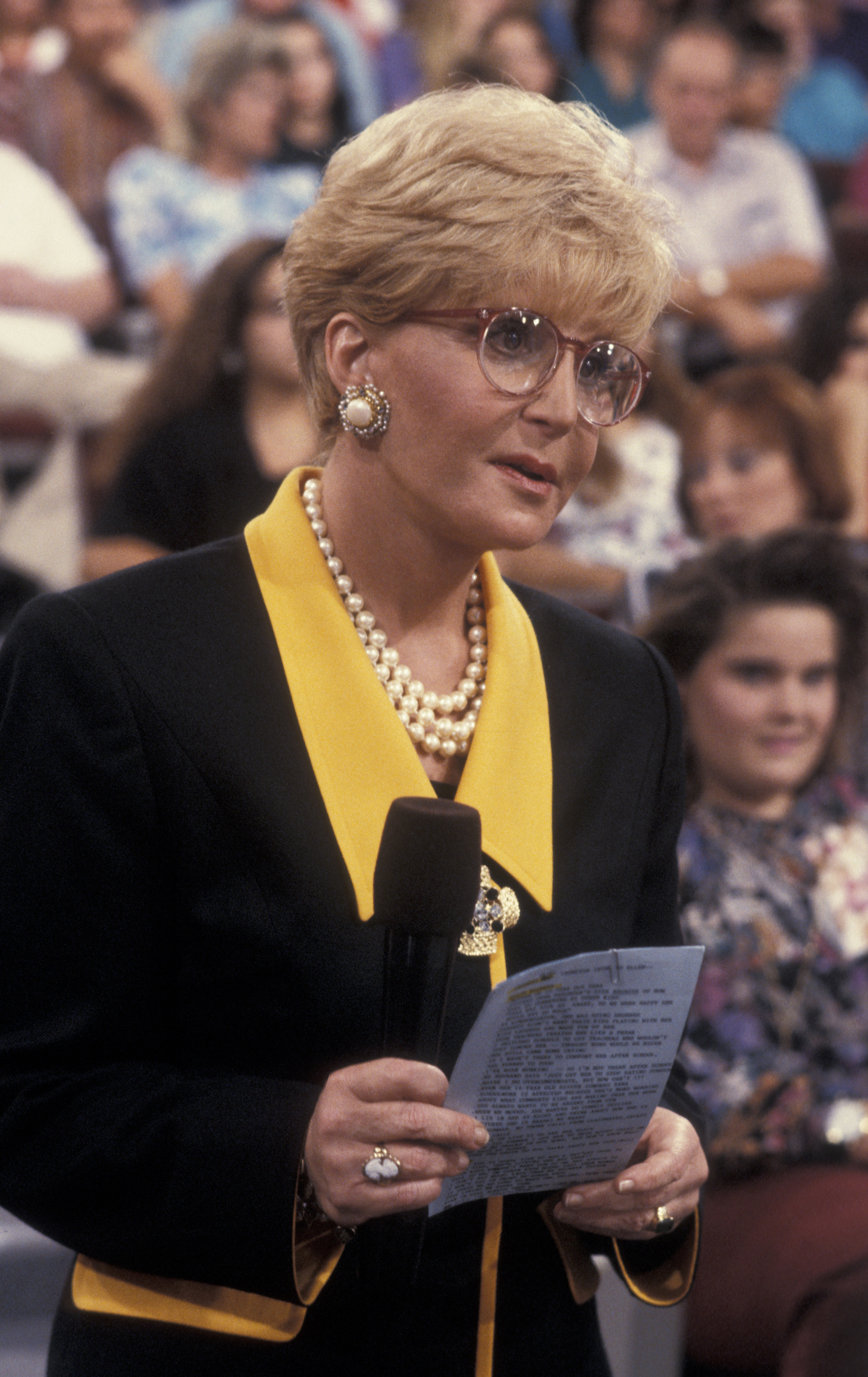 Sally Jessy Raphael addresses her studio audience during the taping of "The Sally Jessy Raphael Show" in Los Angeles on September 9, 1991. Dressed in a black and yellow suit with pearl accessories, she holds cue cards while wearing her signature red-rimmed glasses.