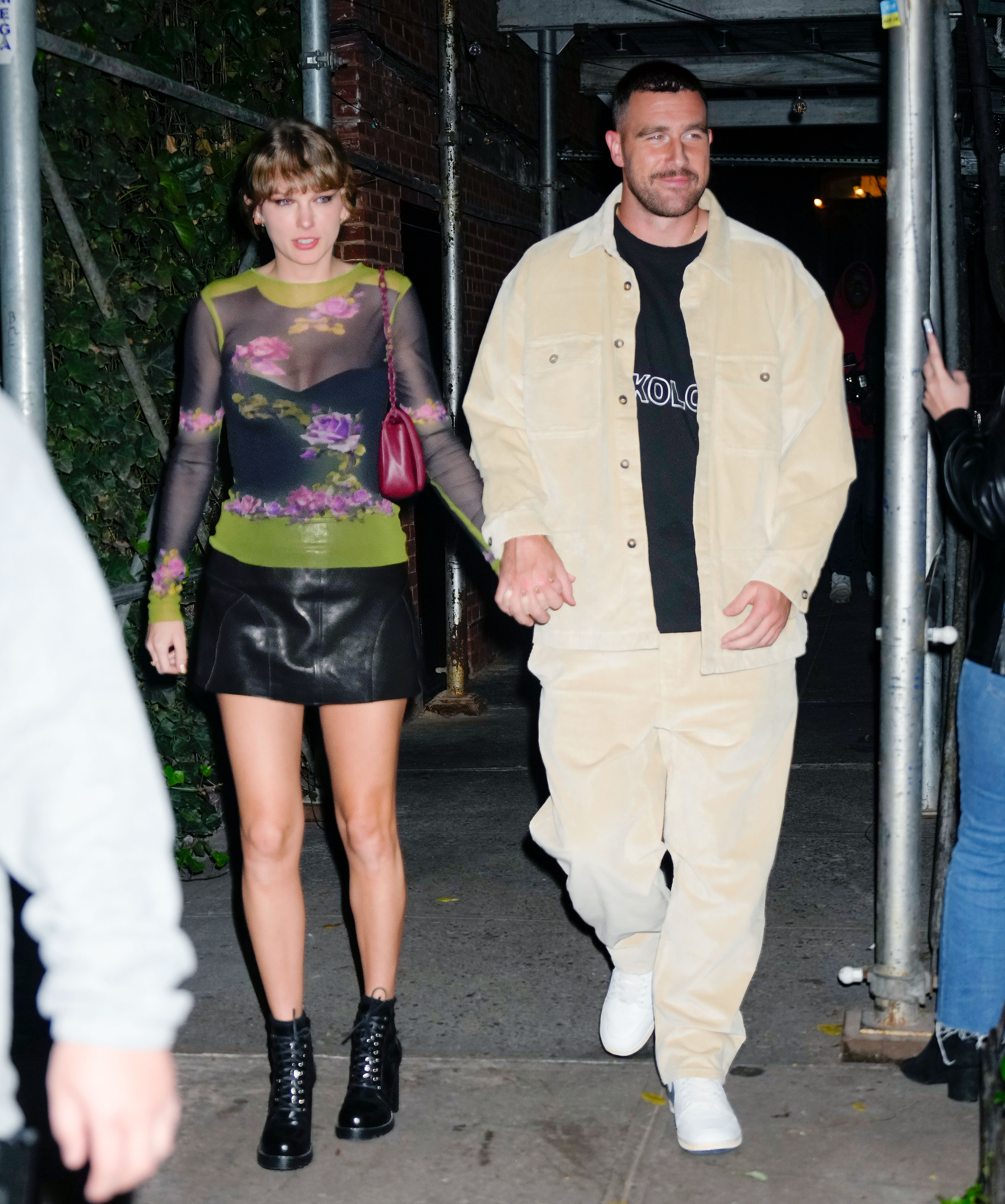 Taylor Swift, wearing a sheer black top with green and purple floral patterns paired with a black leather mini skirt and lace-up boots, holds hands with Travis Kelce, dressed in a beige jacket and pants over a black T-shirt, as they leave Waverly Inn after dinner on October 15, 2023, in New York City.