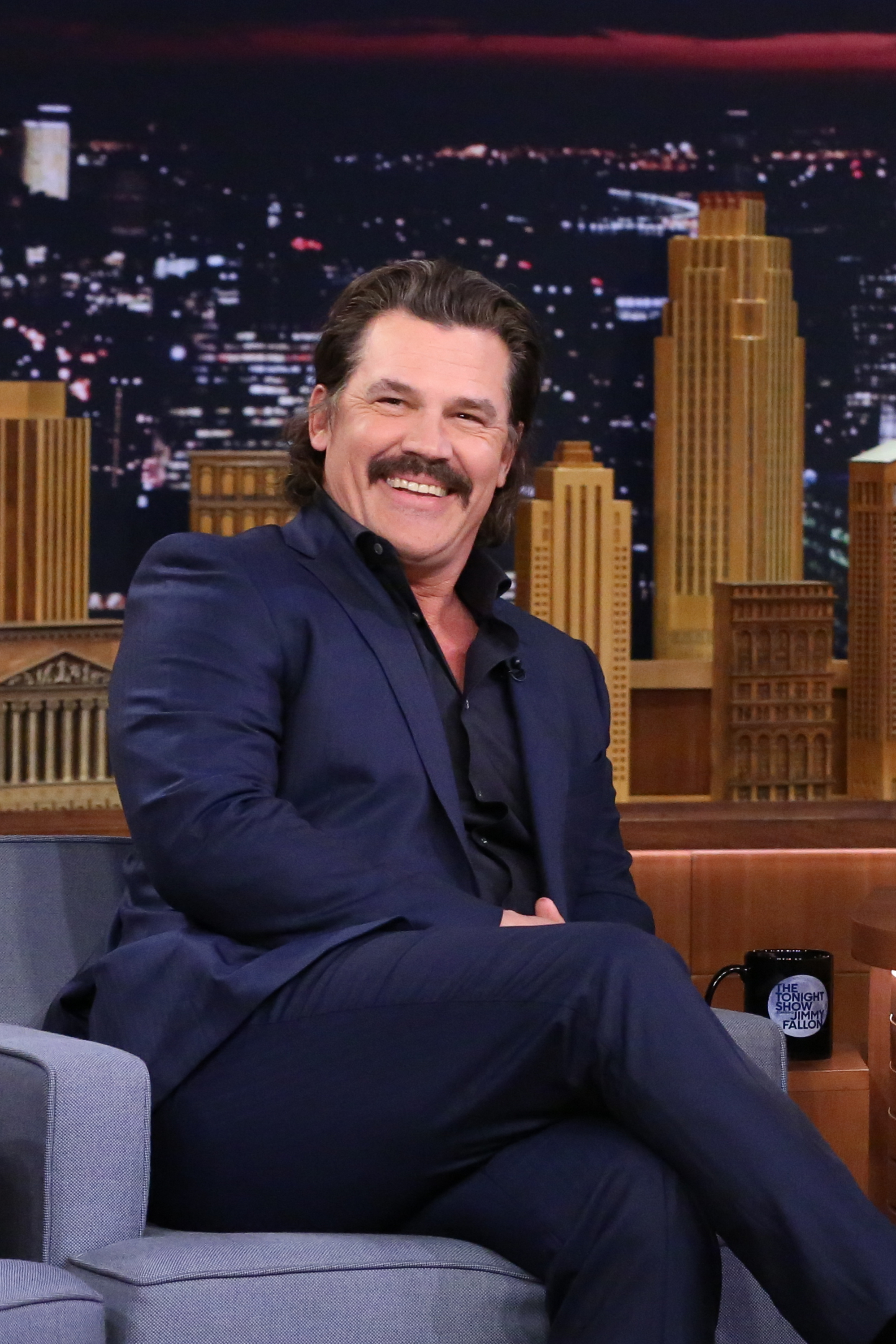 Seated in a navy suit with a dark shirt, he appears at "The Tonight Show Starring Jimmy Fallon," with longer hair and a full mustache — a dramatic shift from his clean-cut looks of earlier years.