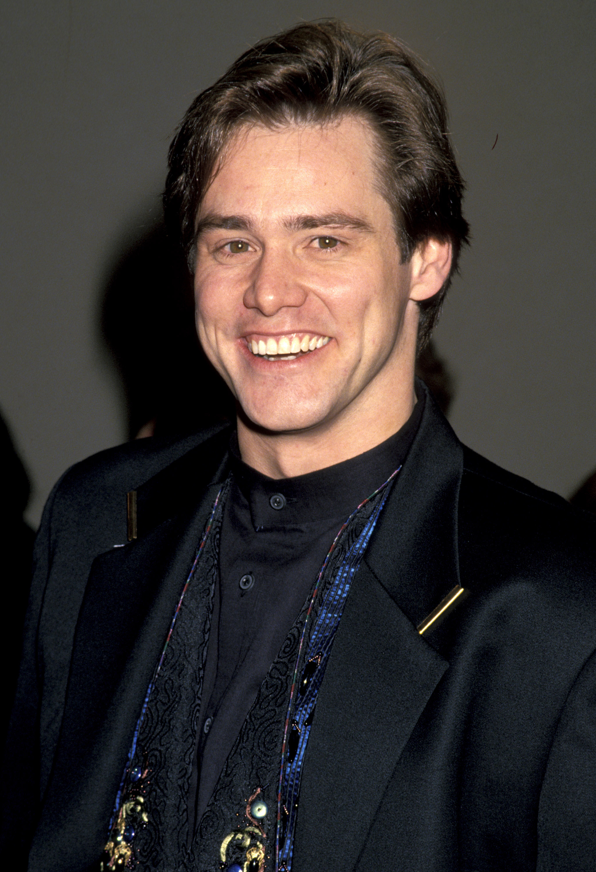 Beaming with a wide, boyish grin, Jim Carrey exudes early-career charm in a sleek black tuxedo layered over an intricately patterned vest at the “Ace Ventura: Pet Detective” premiere. His neatly styled hair and playful confidence reflect a rising star stepping into the spotlight with unmistakable presence.
