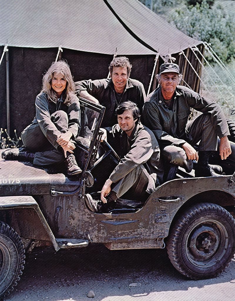 American actor, director and writer Alan Alda in the driving seat of a jeep, surrounded by Loretta Swit and other cast members of the hit television show M.A.S.H, in costume as members of a US Army medical corp. | Getty Images