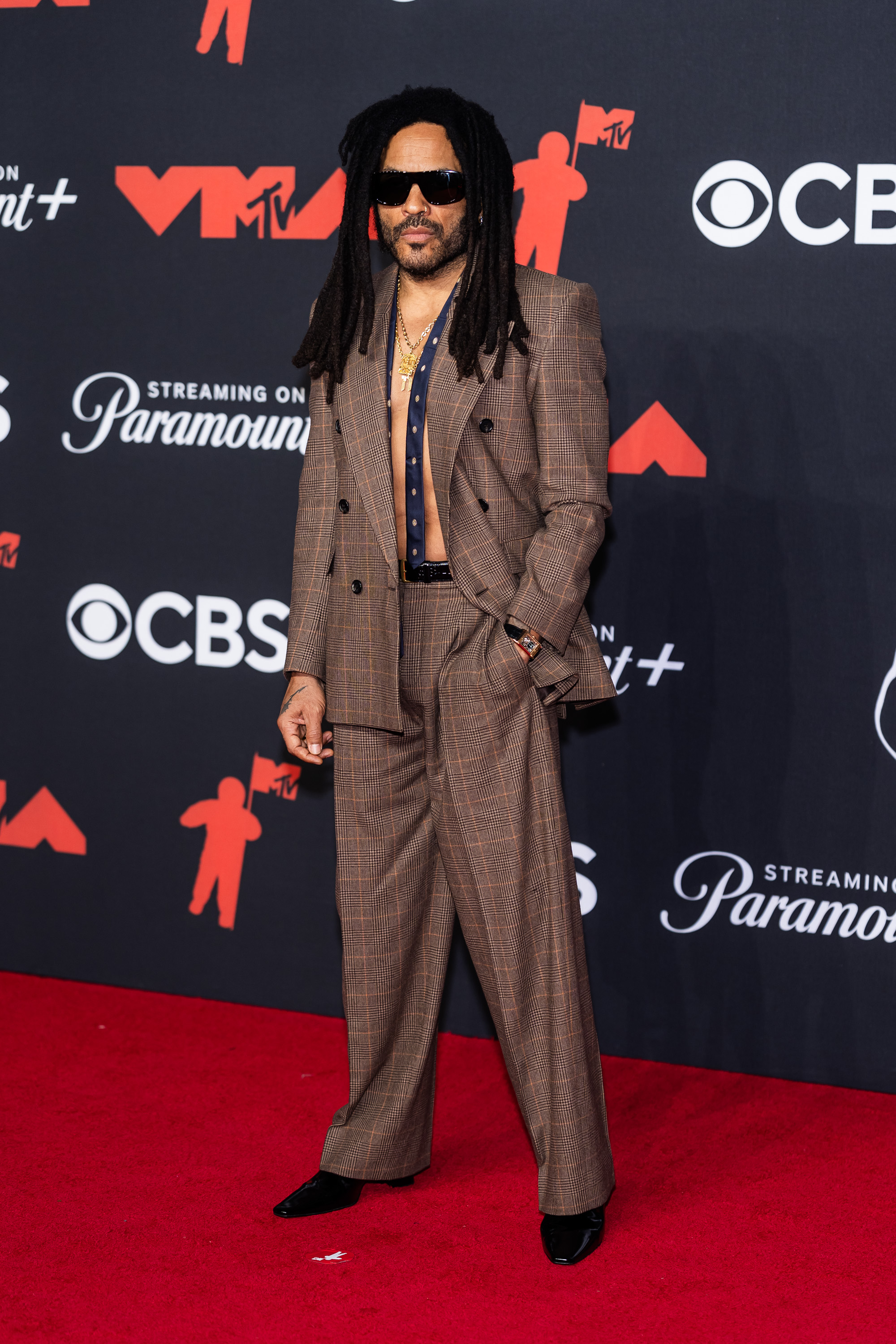 Lenny Kravitz arrives at the 2025 VMAs in a brown checked suit, shirtless beneath the jacket, and accessorized with layered chains and dark sunglasses.