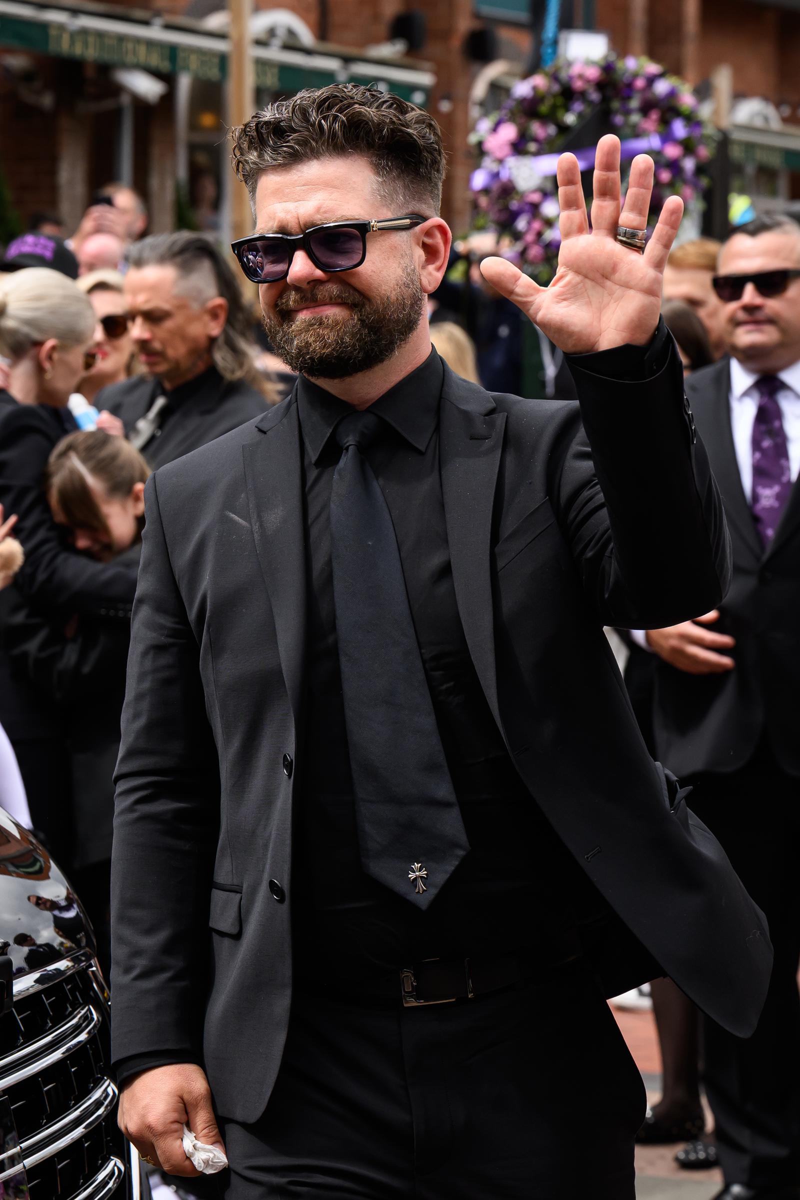 Jack Osbourne raises a hand in acknowledgment as he attends a memorial event honouring his father Ozzy, his expression marked by visible emotion.