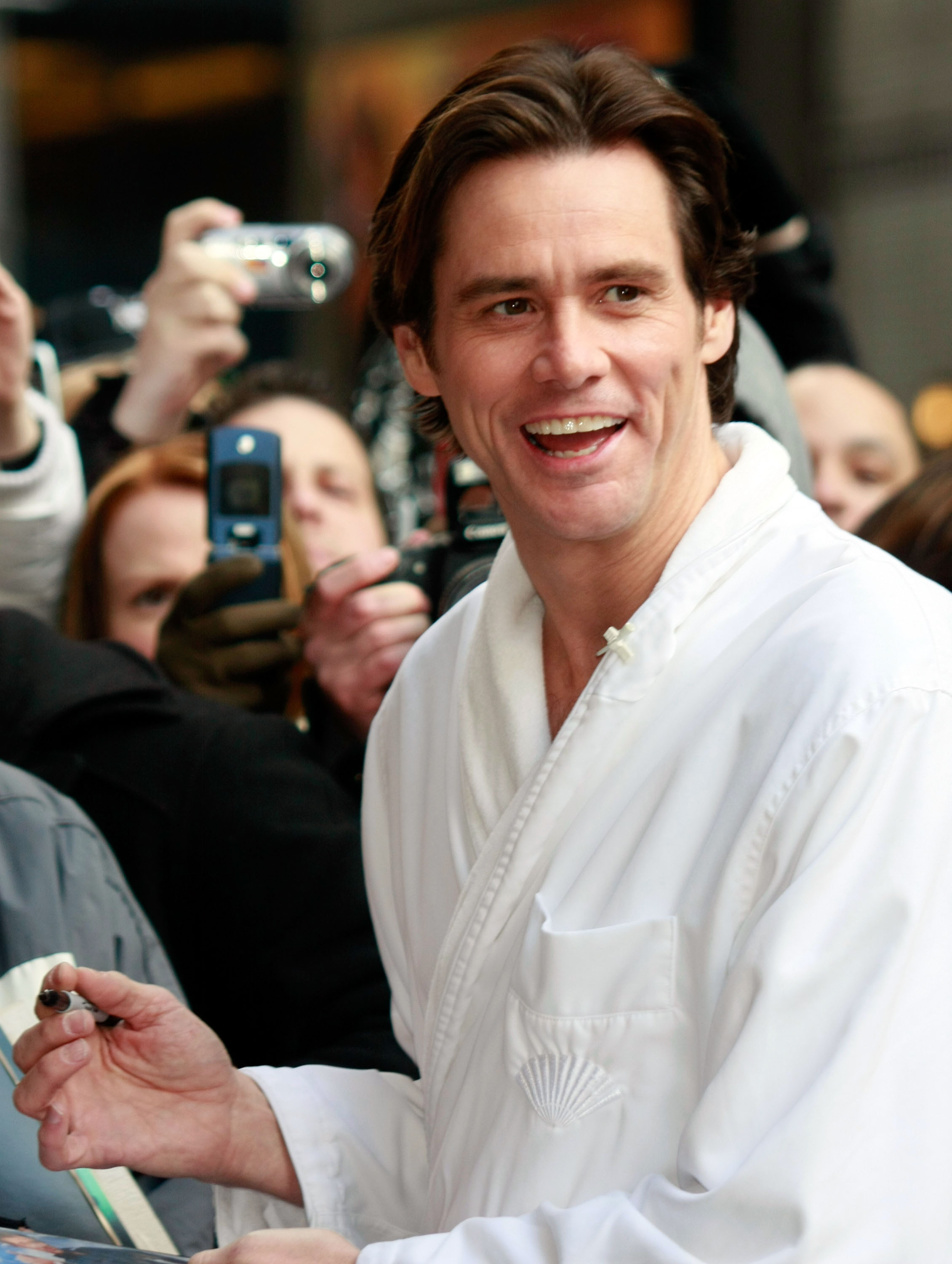 The photo shows actor Jim Carrey smiling brightly as he greets fans outside the Ed Sullivan Theater while visiting the "Late Show with David Letterman." Dressed in a white robe, he appears energetic and playful, signing autographs amid a crowd of admirers. The lively atmosphere captures his charismatic and humorous personality.