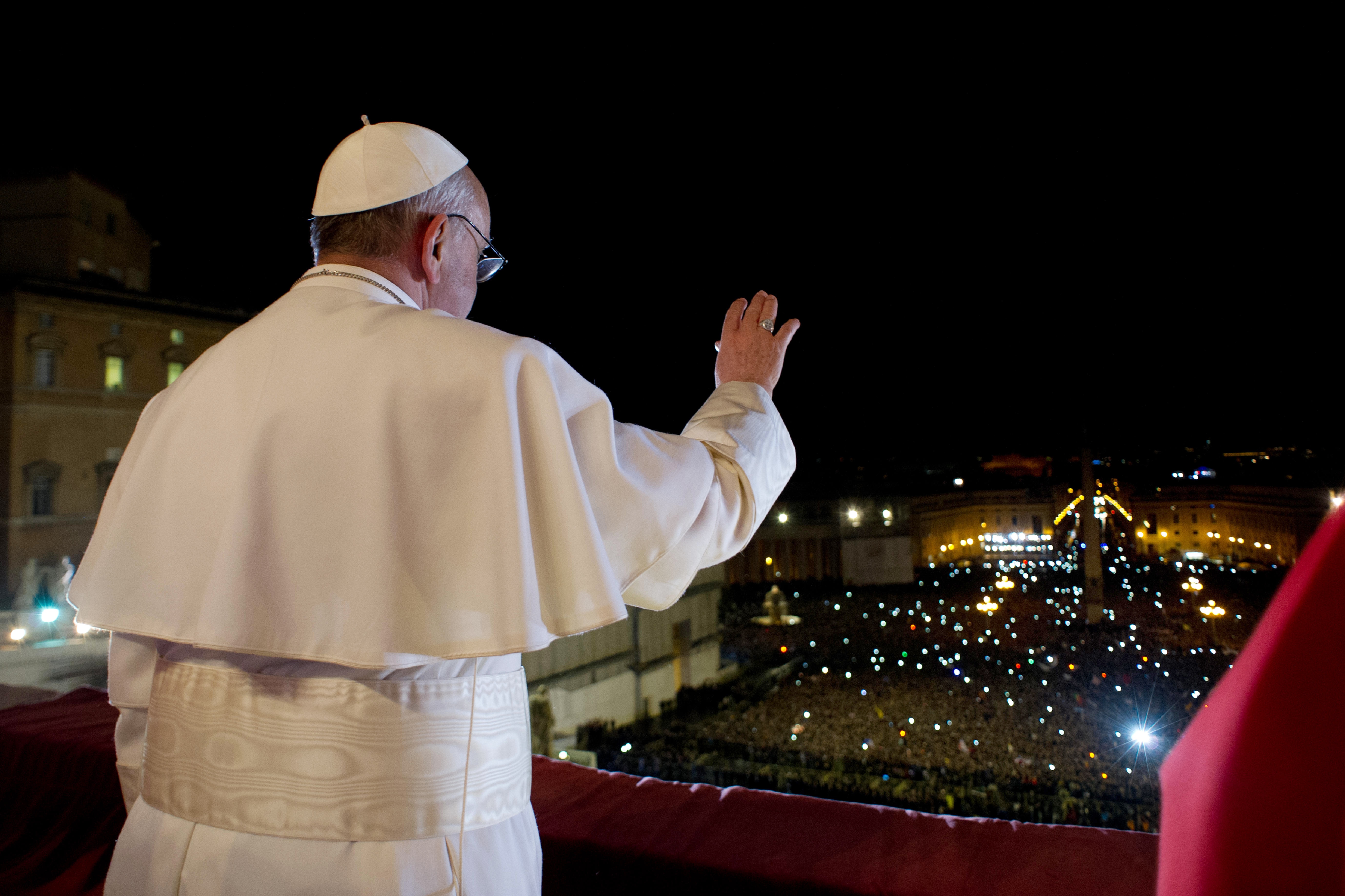 Pope Francis stands in silhouette against the night sky, raising his hand in blessing as thousands of lights sparkle across St. Peter's Square below