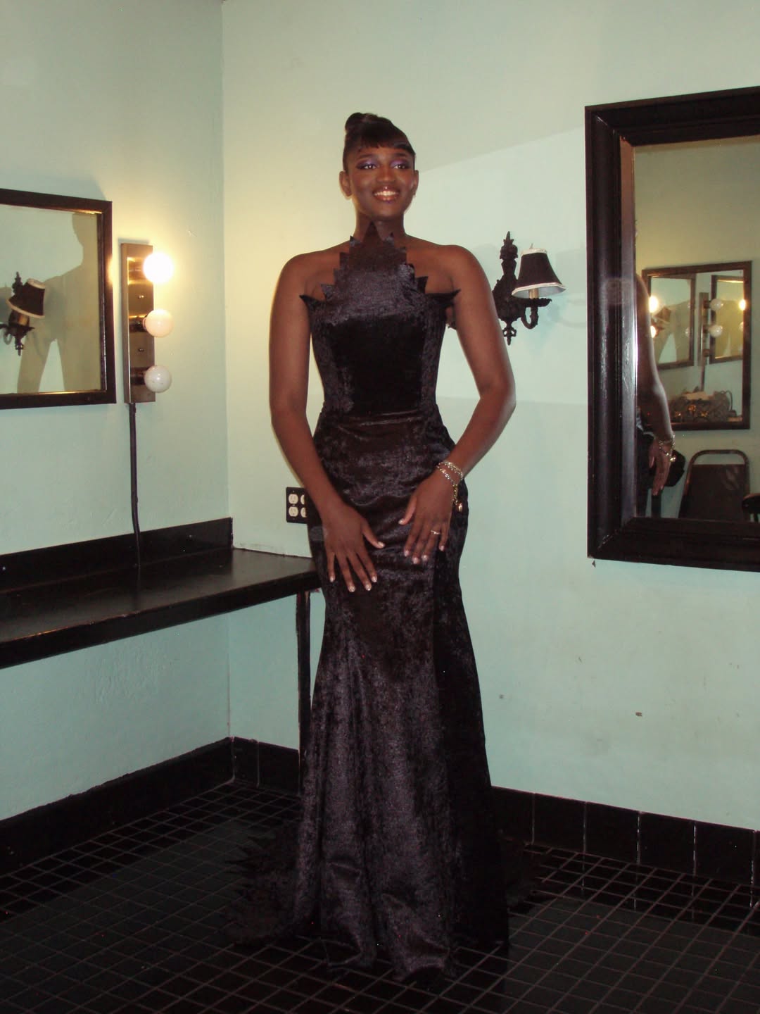Zaya Wade smiles while posing in a sculpted black gown, standing in a dressing room framed by mirrors, vanity lights, and vintage-style wall fixtures. | Source: Instagram/zayawade