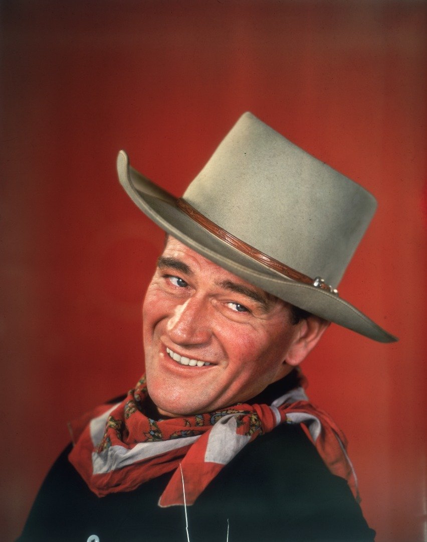 Studio headshot portrait of John Wayne smiling in front of a red background, dressed in Western garb, with his head turned to the side. | Source: Getty Images