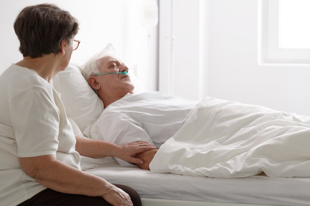 Senior man in a coma laying on hospital bed and his wife sitting next to him. | Photo: Shutterstock