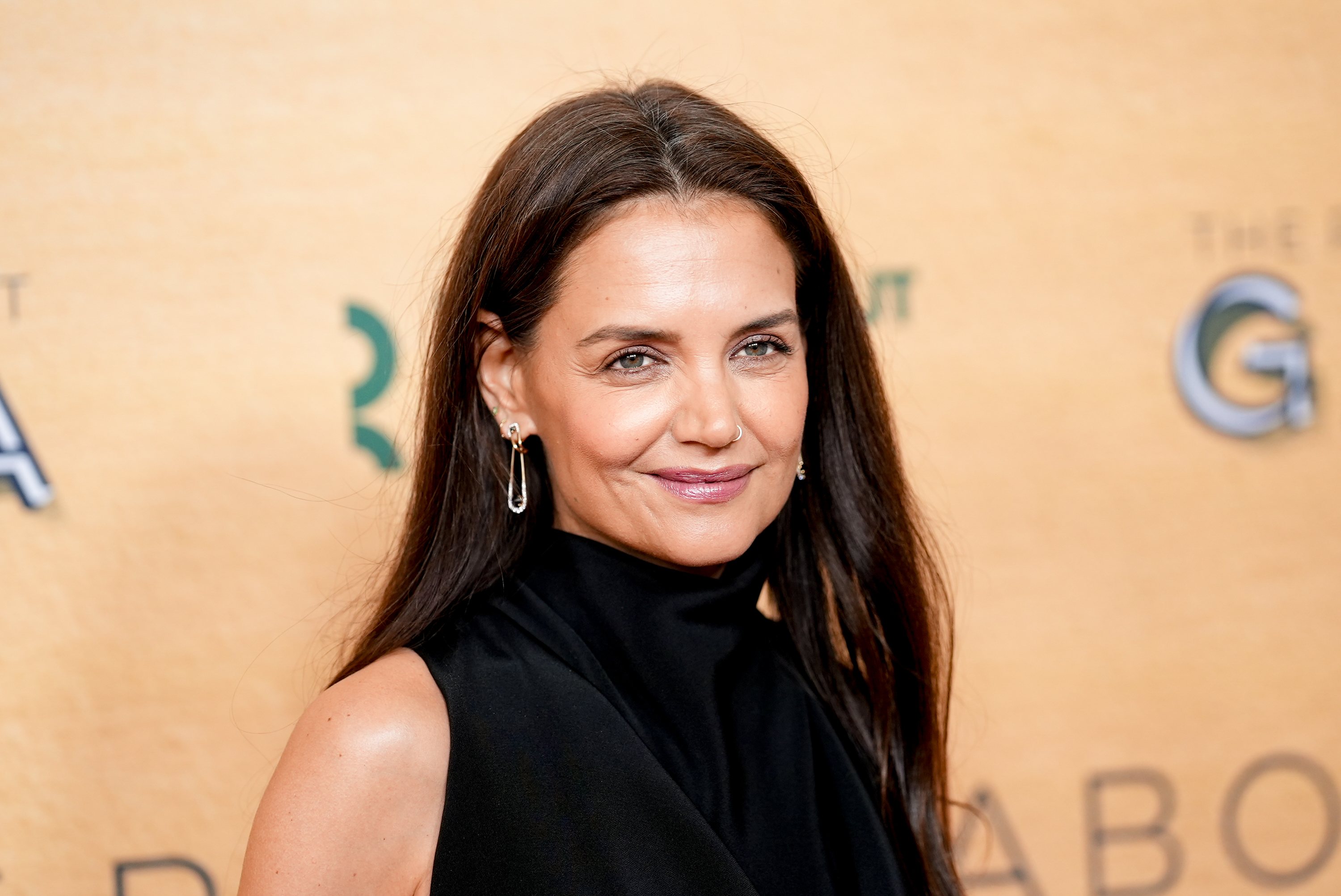 Katie Holmes delivered timeless red-carpet polish in a sleek black halter look accented by delicate jewelry and glossy brunette hair. Against the warm golden backdrop, the actress exuded understated confidence with a modern Hollywood touch.