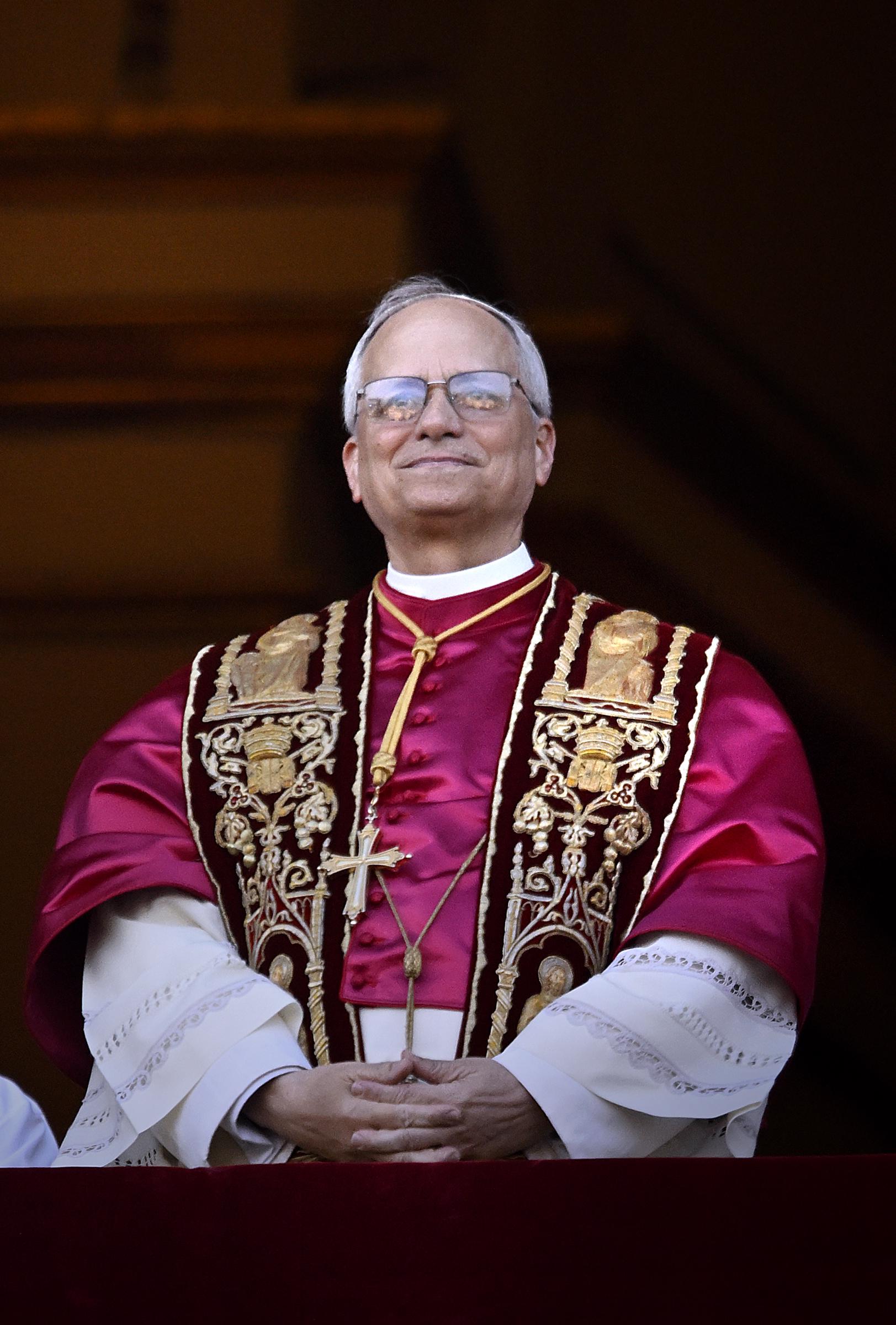 Pope Leo XIV stands with a quiet smile, hands gently clasped over crimson vestments embroidered with gold, his gaze steady beneath gleaming glasses