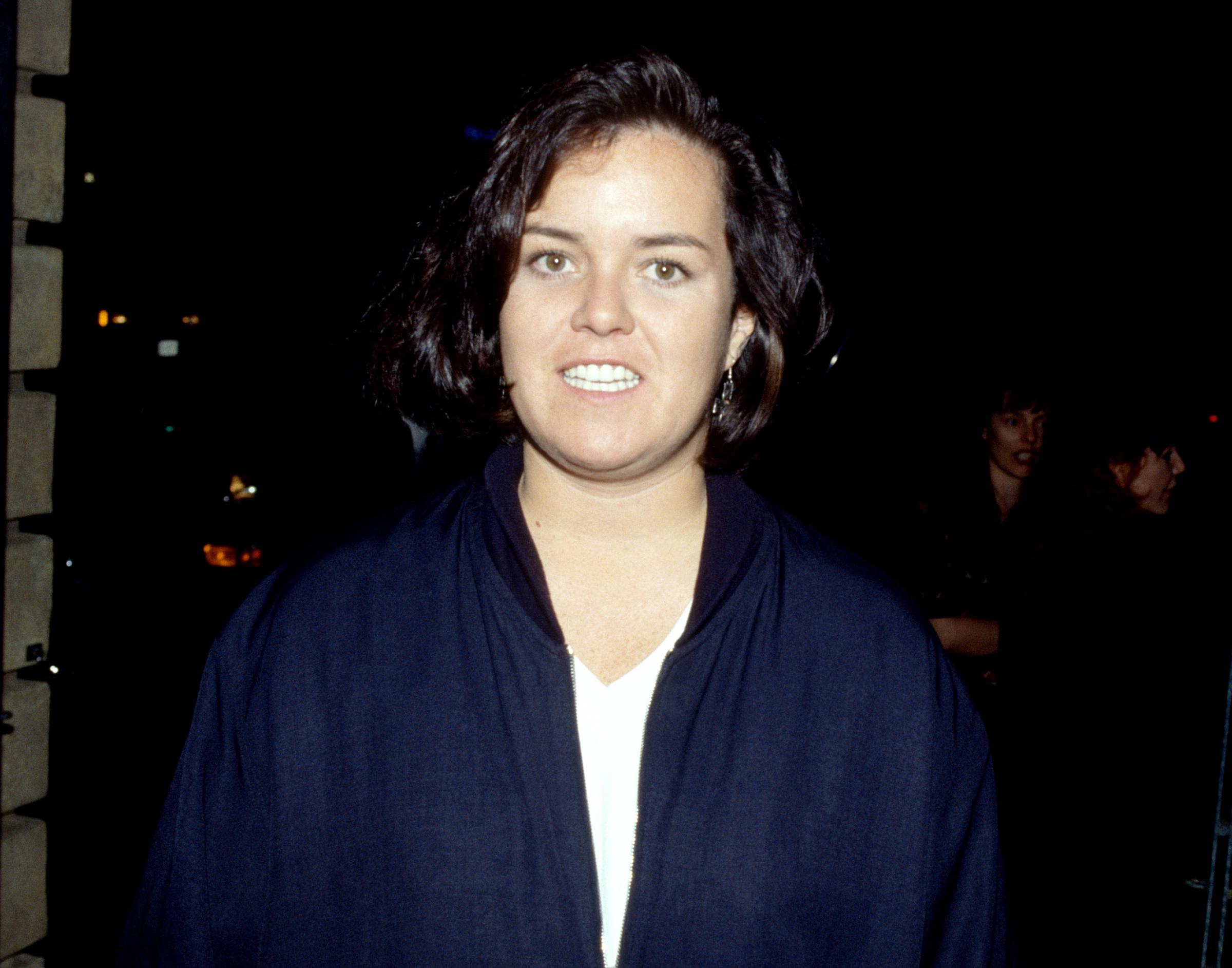 Rosie O'Donnell poses for a portrait in Los Angeles, smiling directly at the camera while standing outdoors at night in a dark oversized jacket against a shadowy city backdrop.
