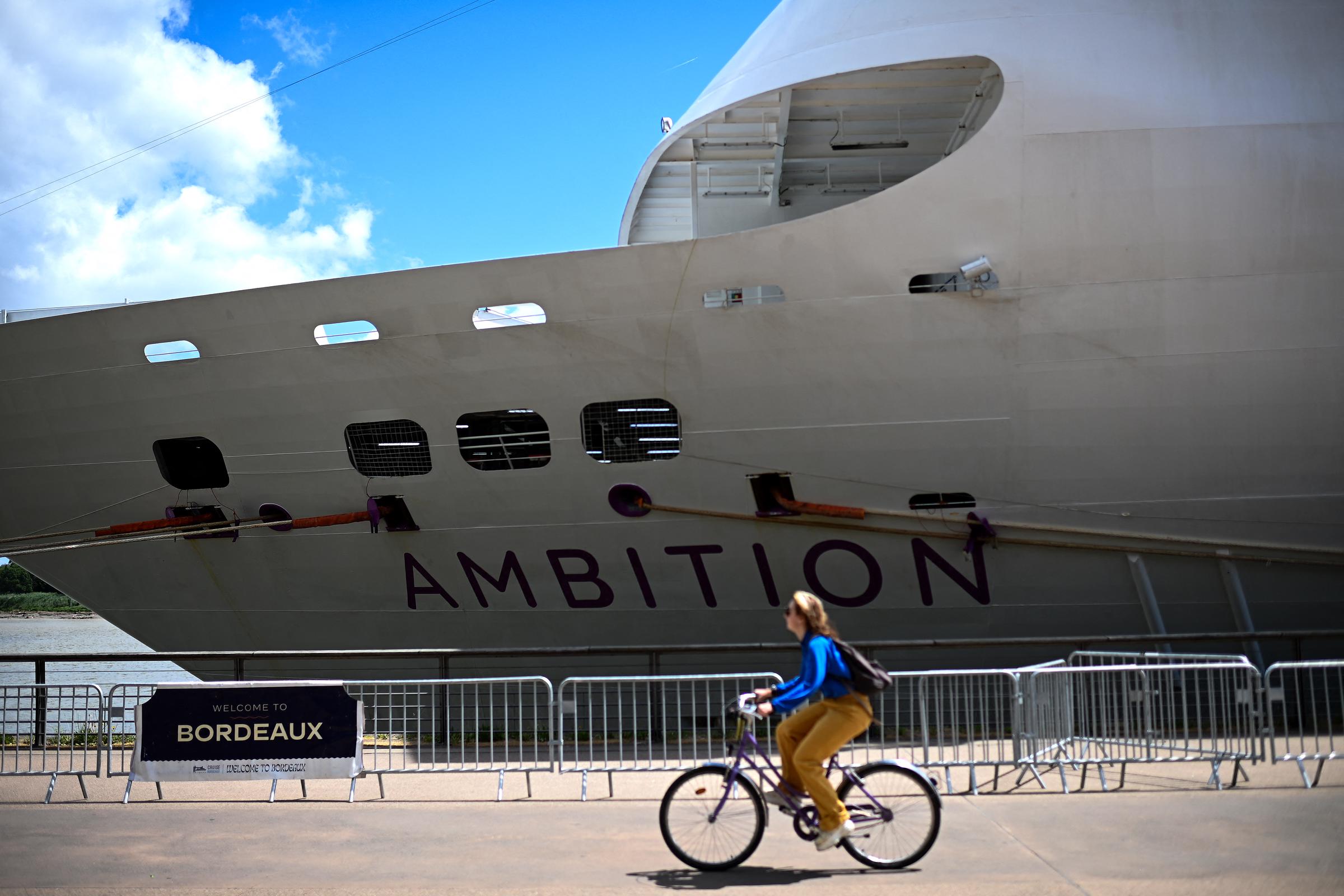 A woman rides a bicycle past the Bahamas-registered cruise ship Ambition after passengers were confined following the outbreak of a gastrointestinal illness onboard, at the Bordeaux port in Bordeaux, southwestern France on May 13, 2026. | Source: Getty Images