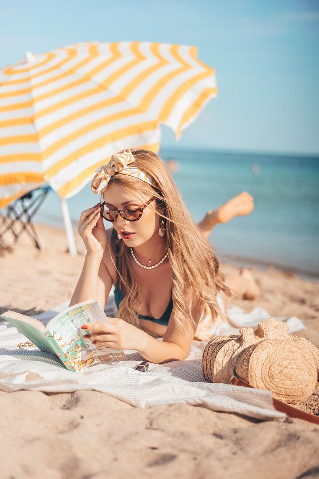 Une jeune femme allongée sur le sable, les lunettes de soleil aux yeux et un magazine dans les mains | Photo : Pexels