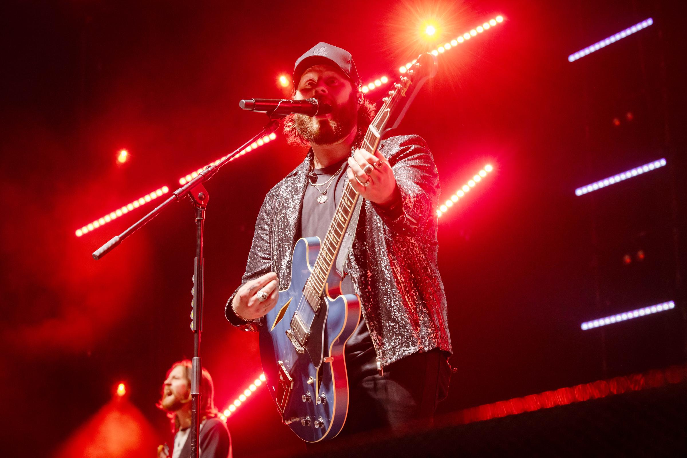Nate Smith performs under fiery red lights, singing and playing a blue guitar while his shimmering jacket catches the glow of the stage.