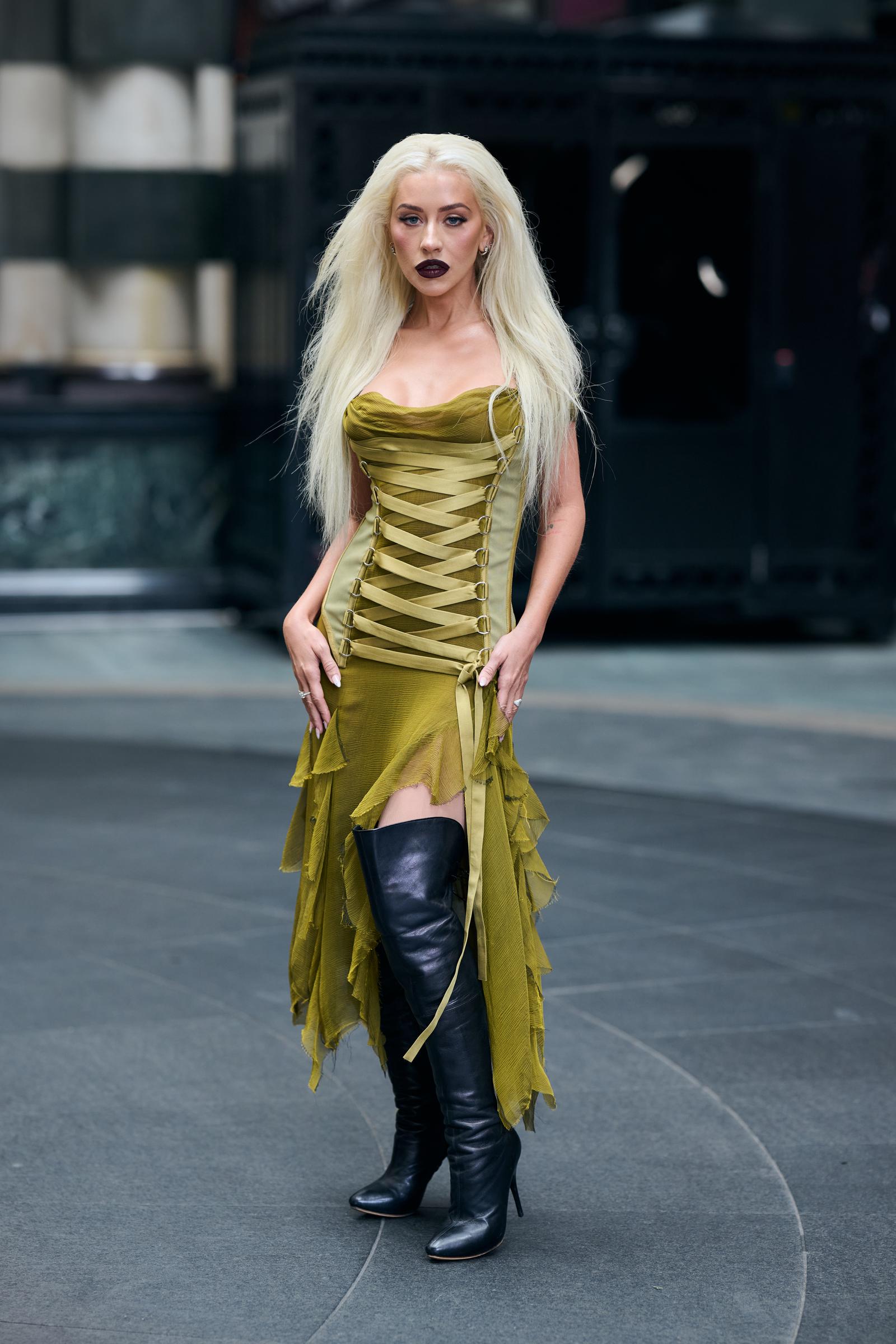 Christina Aguilera attends the photocall for "Burlesque: The Musical" at The Savoy Theatre in London on July 21, 2025. She poses in an olive green corset dress with ribbon lace-up detailing and an asymmetrical hemline, paired with black knee-high boots and sleek, platinum blonde hair cascading down her shoulders.