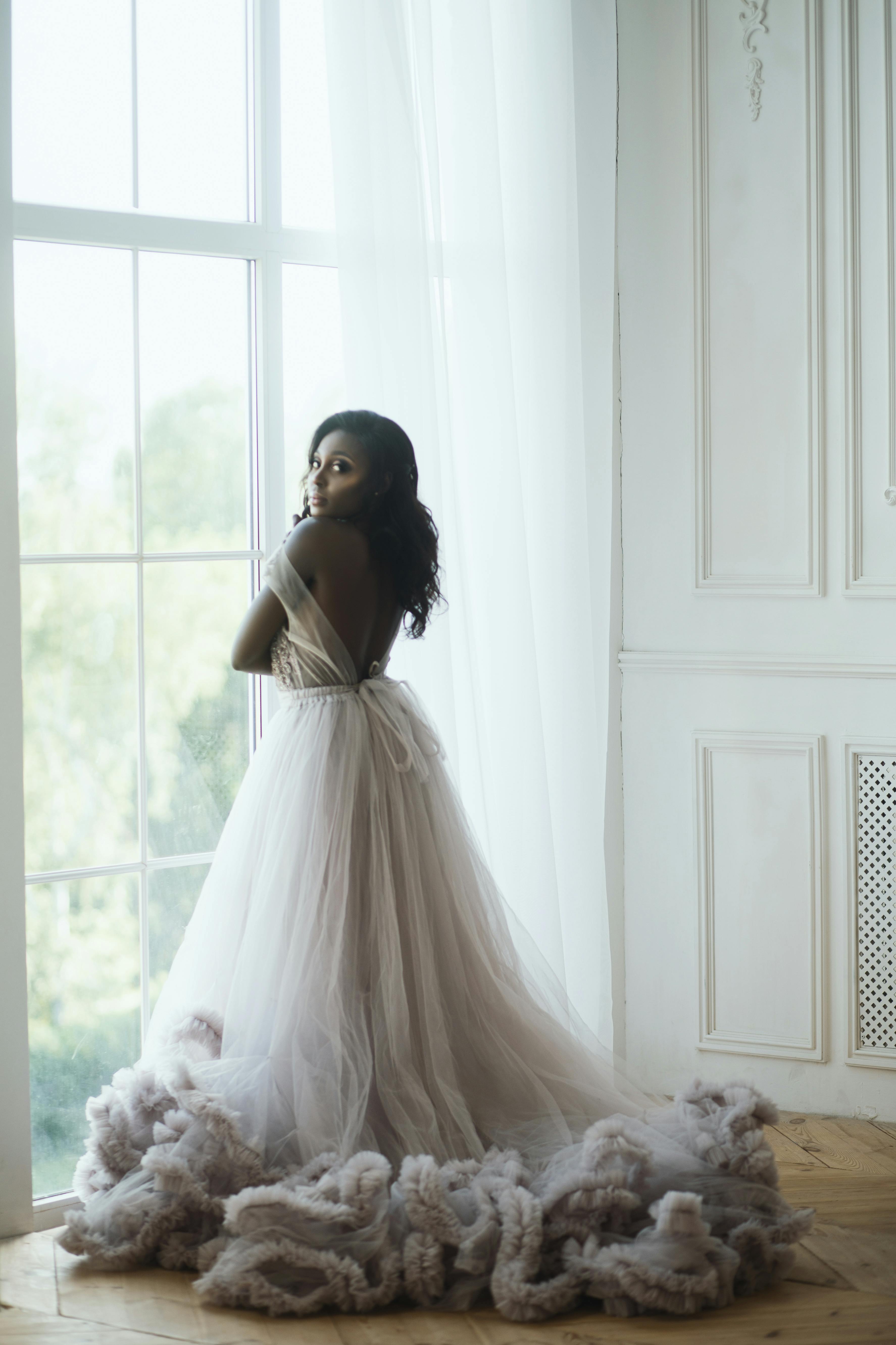 For illustration purposes only. Woman standing at a window in a wedding dress looks at the camera | Source: Pexels