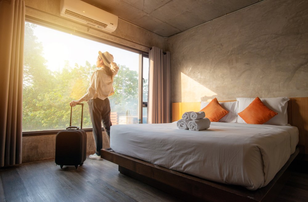 Portrait of tourist woman standing nearly window, looking to beautiful view with her luggage in hotel bedroom after check-in. Conceptual of travel and vacation | Photo: Shutterstock