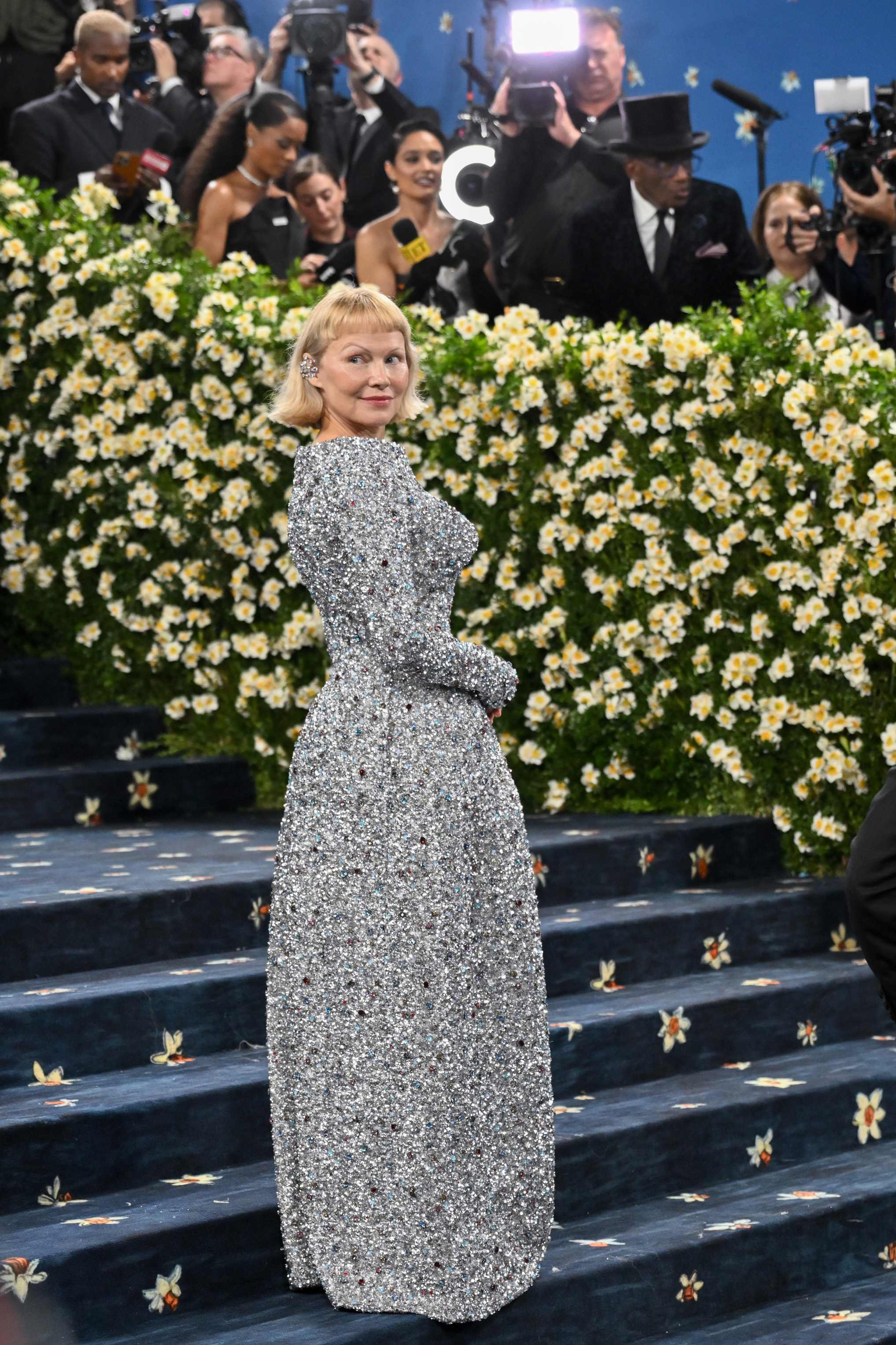 Pamela Anderson turns toward photographers as she poses on the iconic Met Gala steps. The backdrop of yellow and white blooms underscores the night's celebration of "Superfine: Tailoring Black Style" at the Metropolitan Museum of Art.