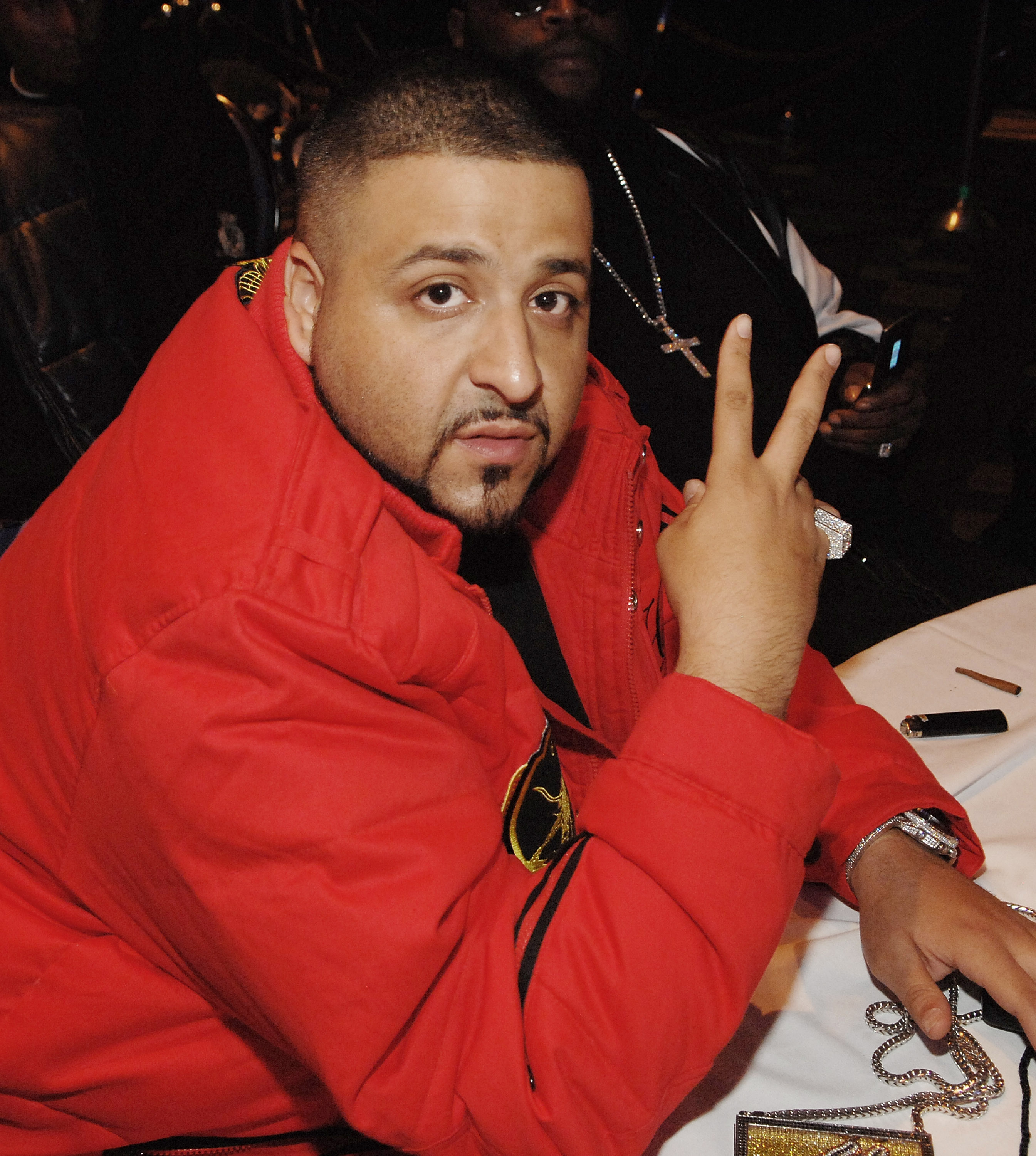 Seated at a table in a bold red jacket, DJ Khaled raises two fingers toward the lens while diamond chains and a pendant rest nearby, adding shine to the dimly lit nightlife setting.