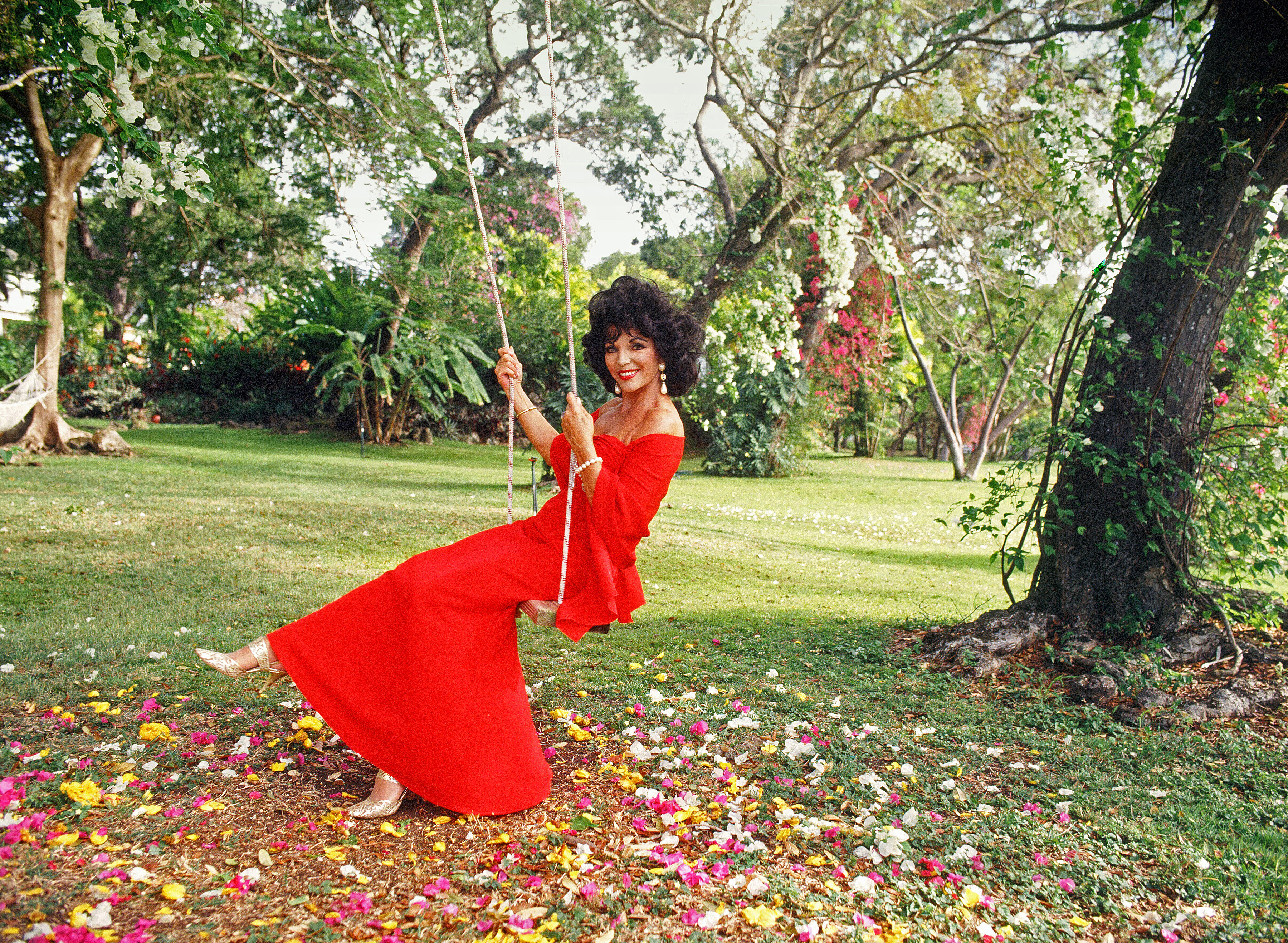 Joan Collins sits on a swing in a red gown in the grounds of the Sugar Bay Resort in Bridgetown, Barbados, February 1994 | Source: Getty Images