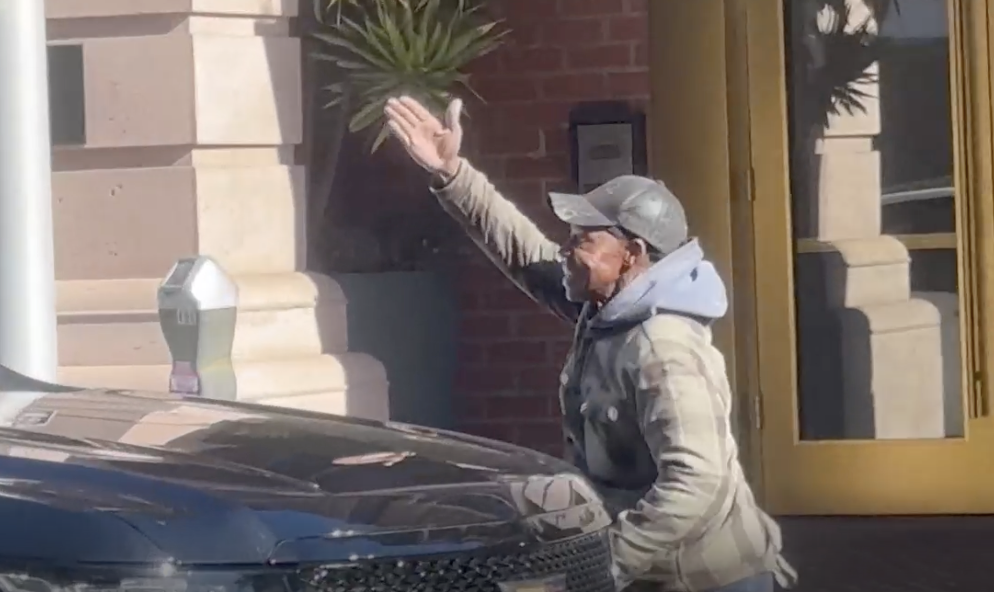A man raised his arm in a blowing kiss gesture while standing beside a black SUV parked outside a building | Source: Youtube/TMZ