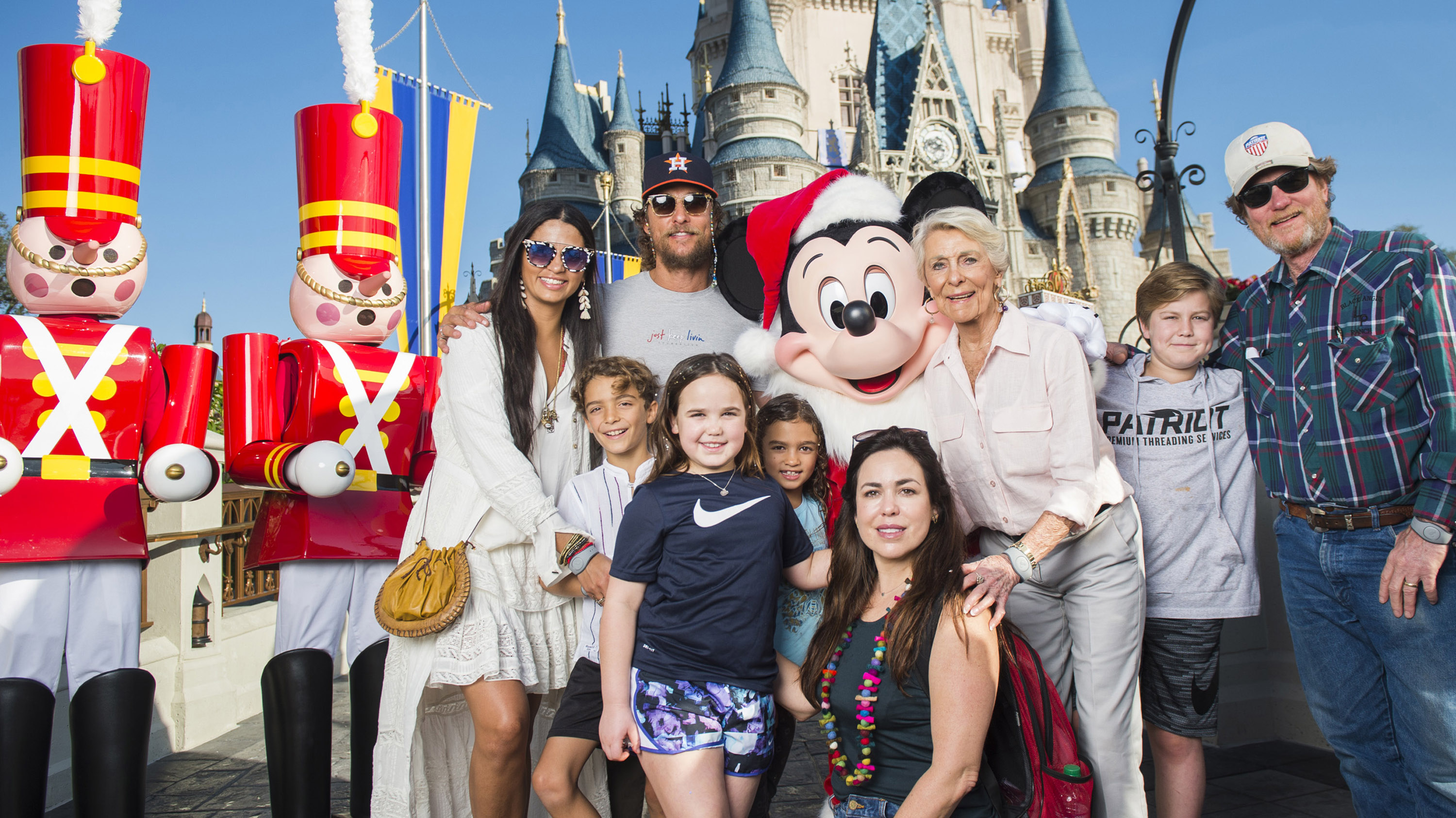 Levi McConaughey and Vida McConaughey stand at the front, smiling among family and park characters. Levi wears a light shirt and shorts, while Vida stands close in a casual outfit among other children. Matthew McConaughey and Camila Alves McConaughey stand behind them with relatives near the castle backdrop.