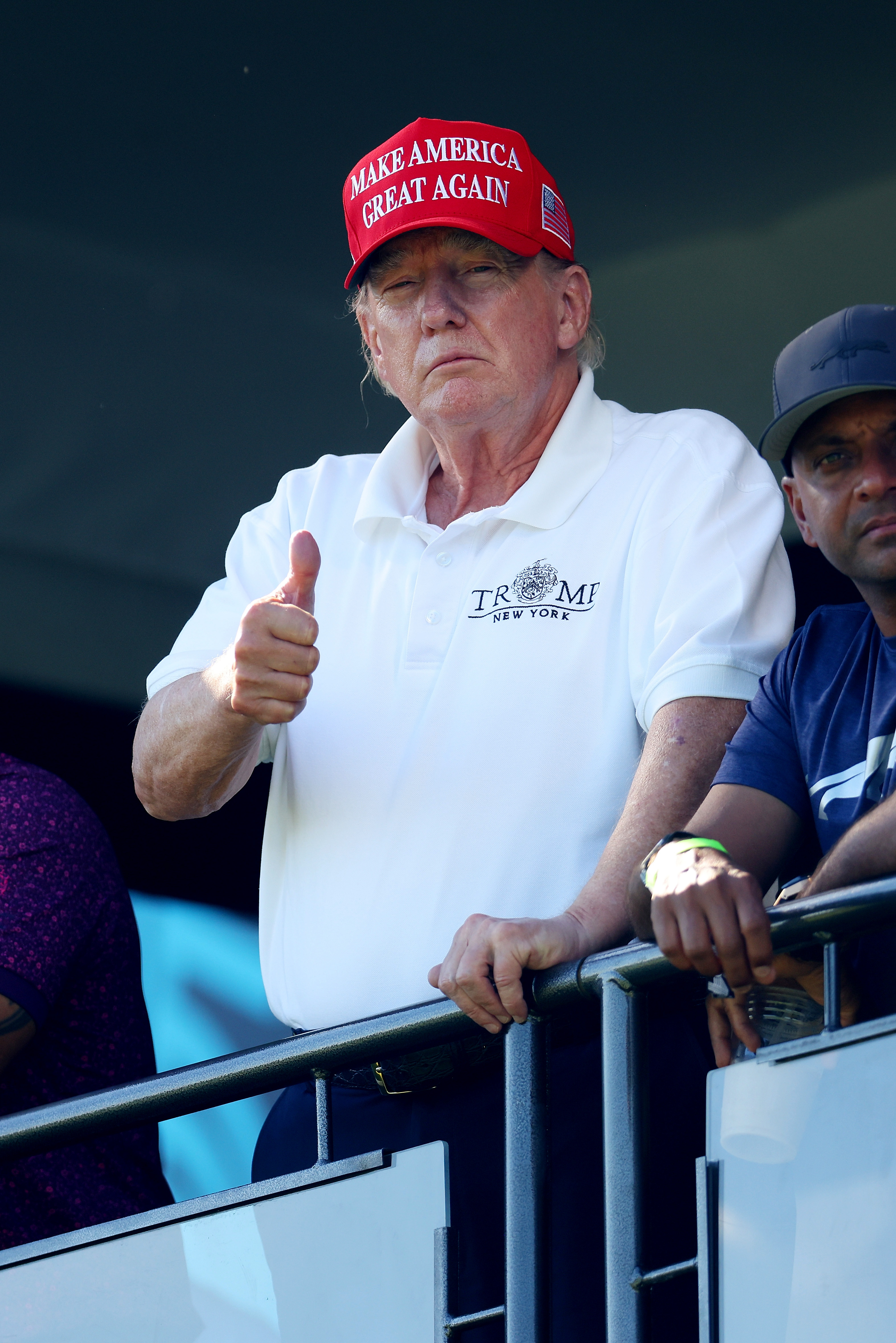 On August 13, Donald Trump stands near a railing at the 18th green, giving a thumbs-up. The redness in his face remains visible, with lighter tones around the edges of his jaw and neck.