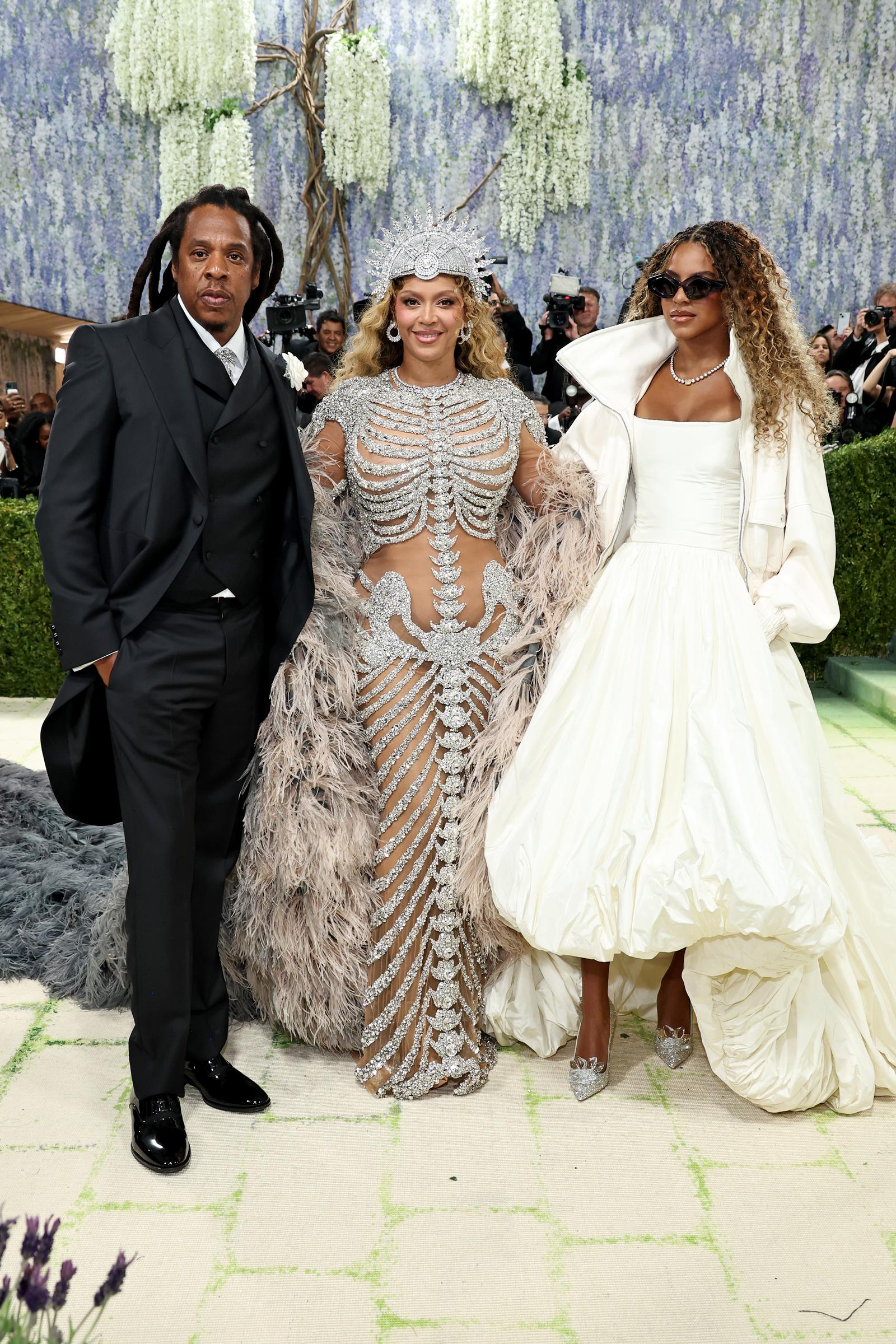 Jay-Z keeps it classic in a sharp black tux, while Beyoncé dazzles in a crystal-covered illusion gown with a striking silver headpiece. Beside them, Blue Ivy Carter adds a modern touch in a voluminous white gown and sleek shades.