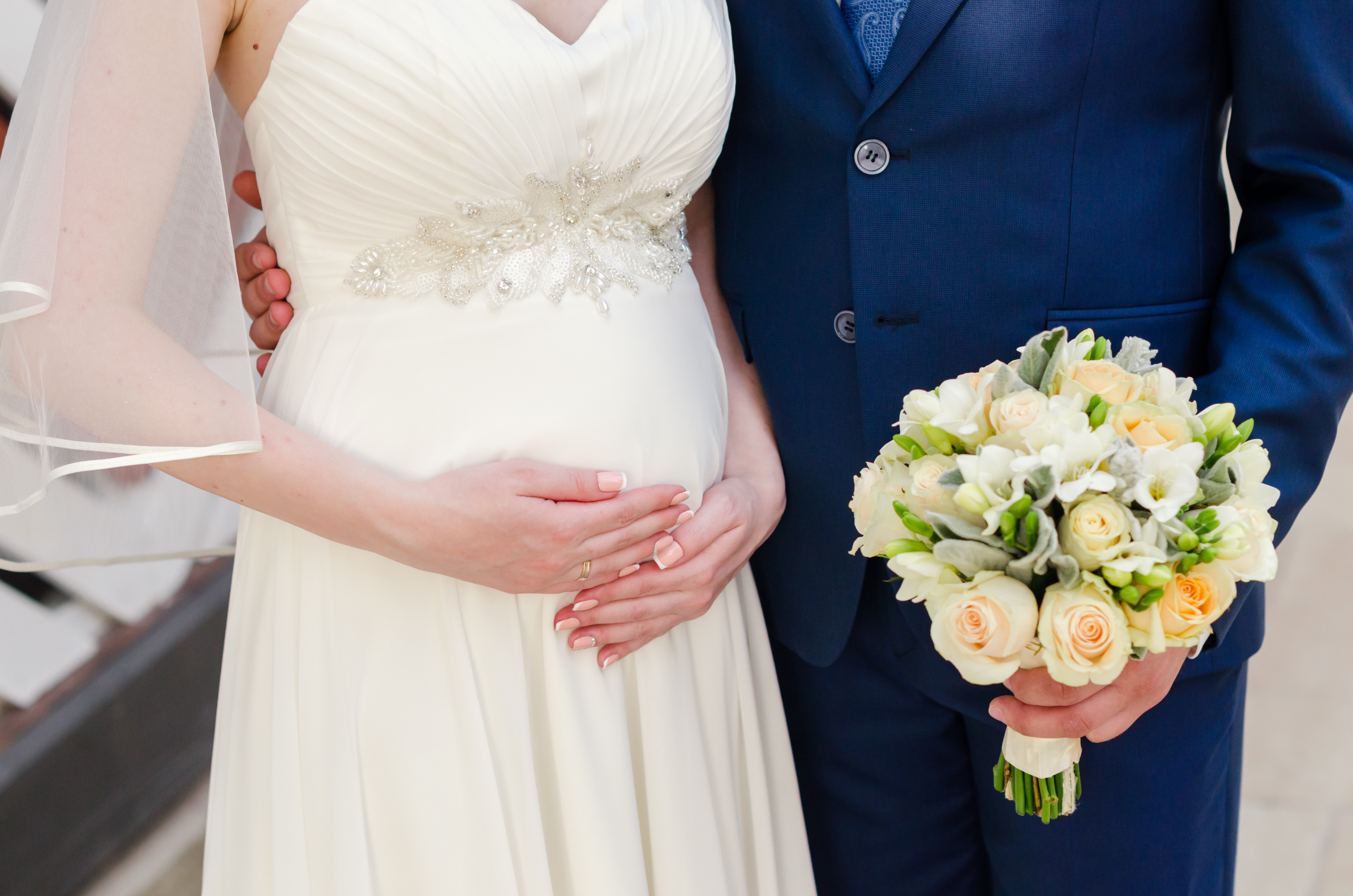 A pregnant bride holds her belly as her groom holds her bouquet | Source: Shutterstock