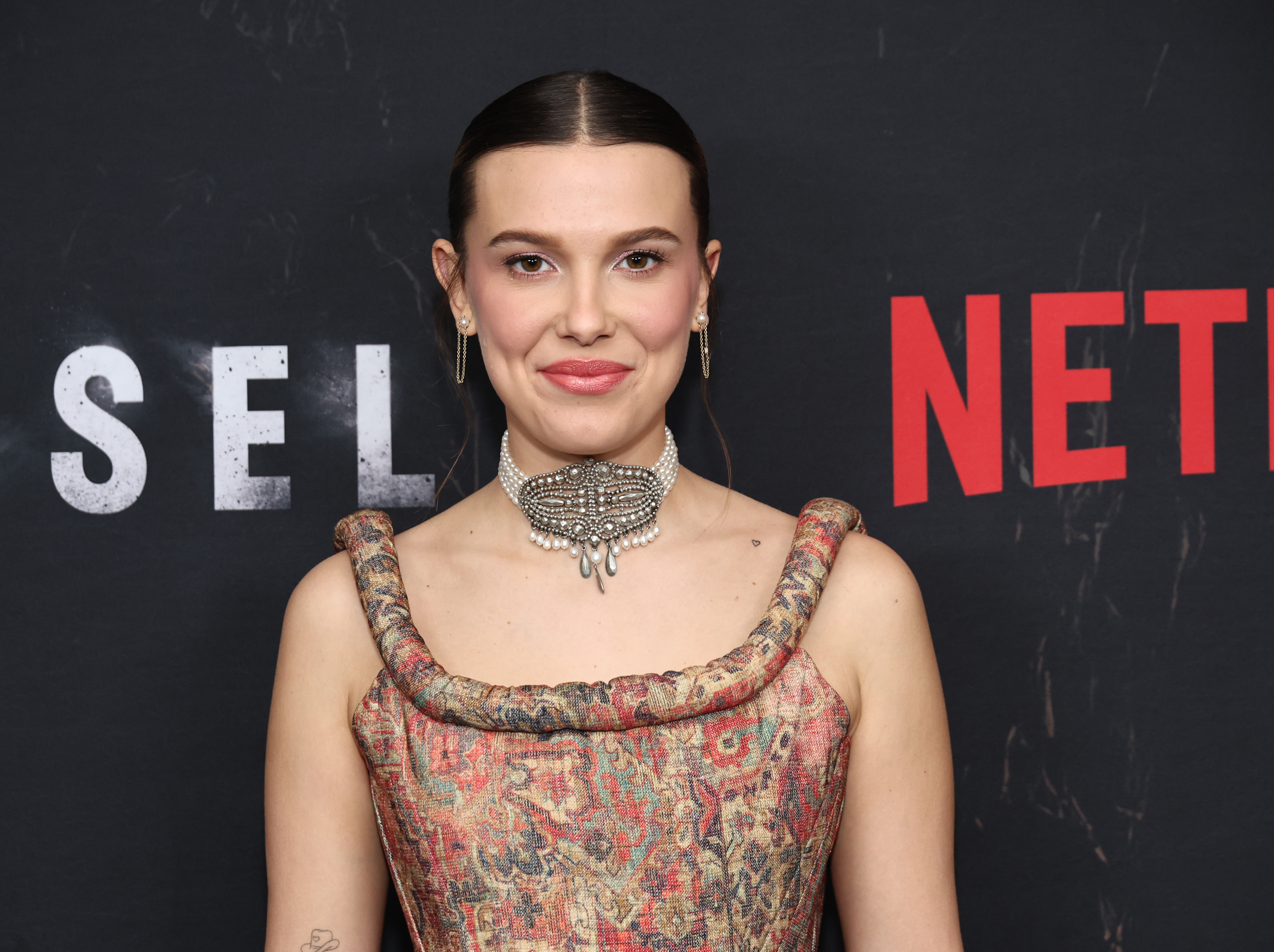 At a Netflix event, Millie Bobby Brown opts for refined drama in a textured corset-style dress. A pearl-adorned choker anchors the look, drawing focus to her poised expression. It's a sophisticated moment that signals confident evolution.