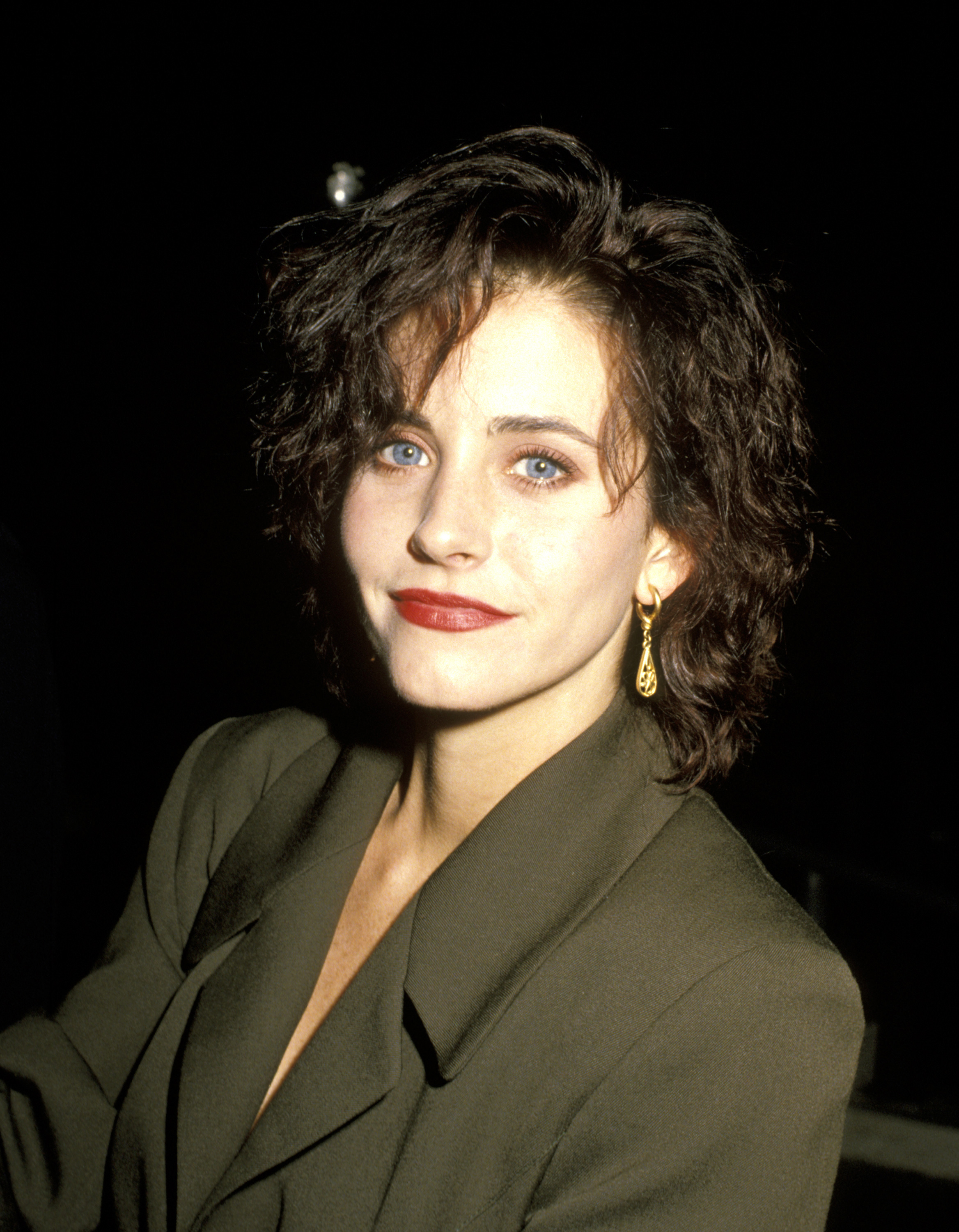 Courteney Cox stuns in a 1990 close-up, highlighting her striking blue eyes, bold red lipstick, and gold dangling earrings, all paired with a stylish green suit.