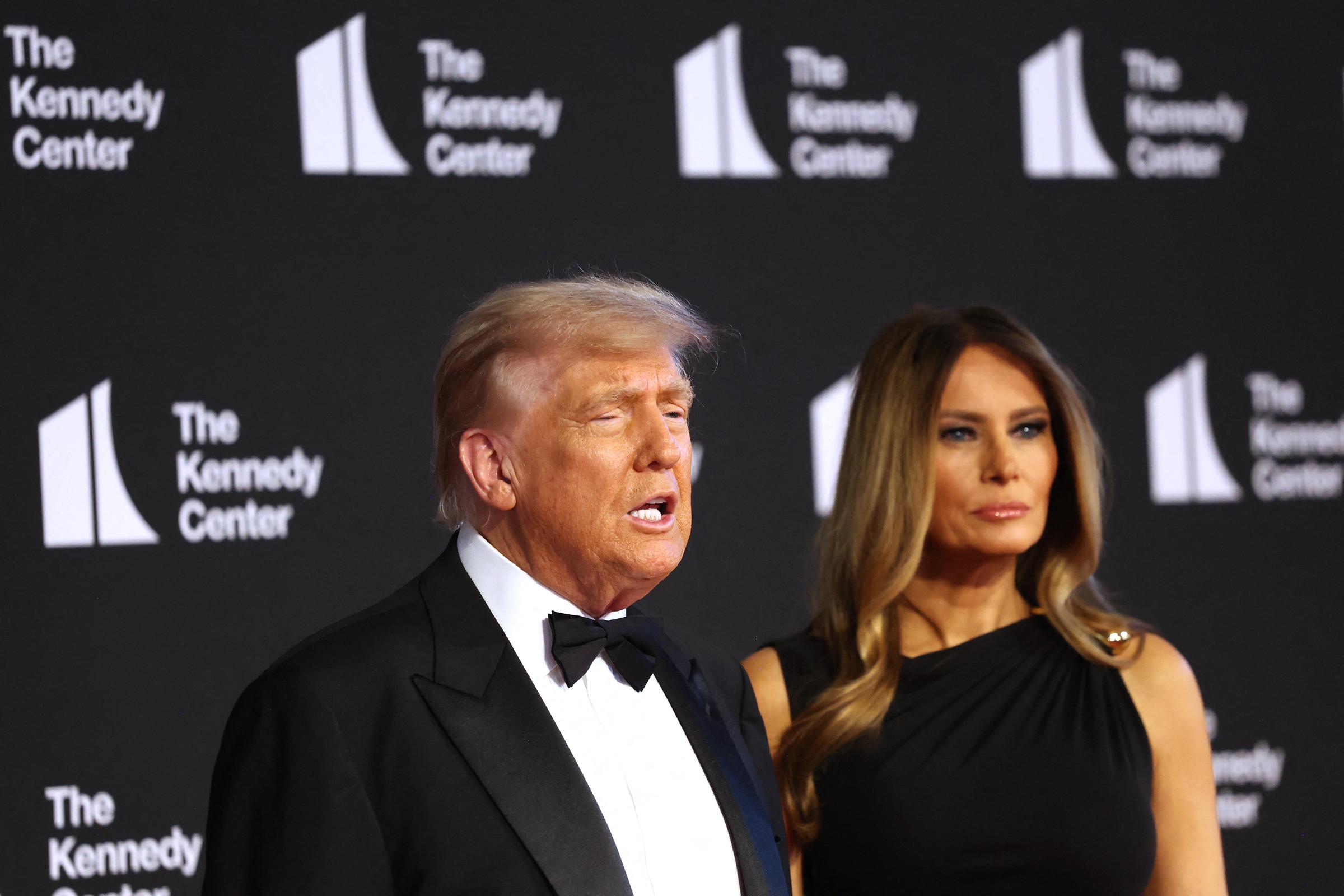 Donald and Melania stand posing for the paparazzi. Donald's hair is light blonde in a swept-back, neatly combed style. Melania's hair is long and softly curled at the ends, featuring a smooth blend of honey blonde and light brown tones with a glossy finish.