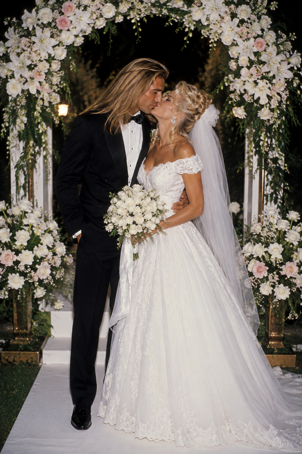 In an AI-generated scene, Fabio and Anna Nicole Smith share a kiss under a lavish white floral arch, dressed in classic wedding attire that feels straight out of a mid-1990s celebrity ceremony. | Source: ChatGPT