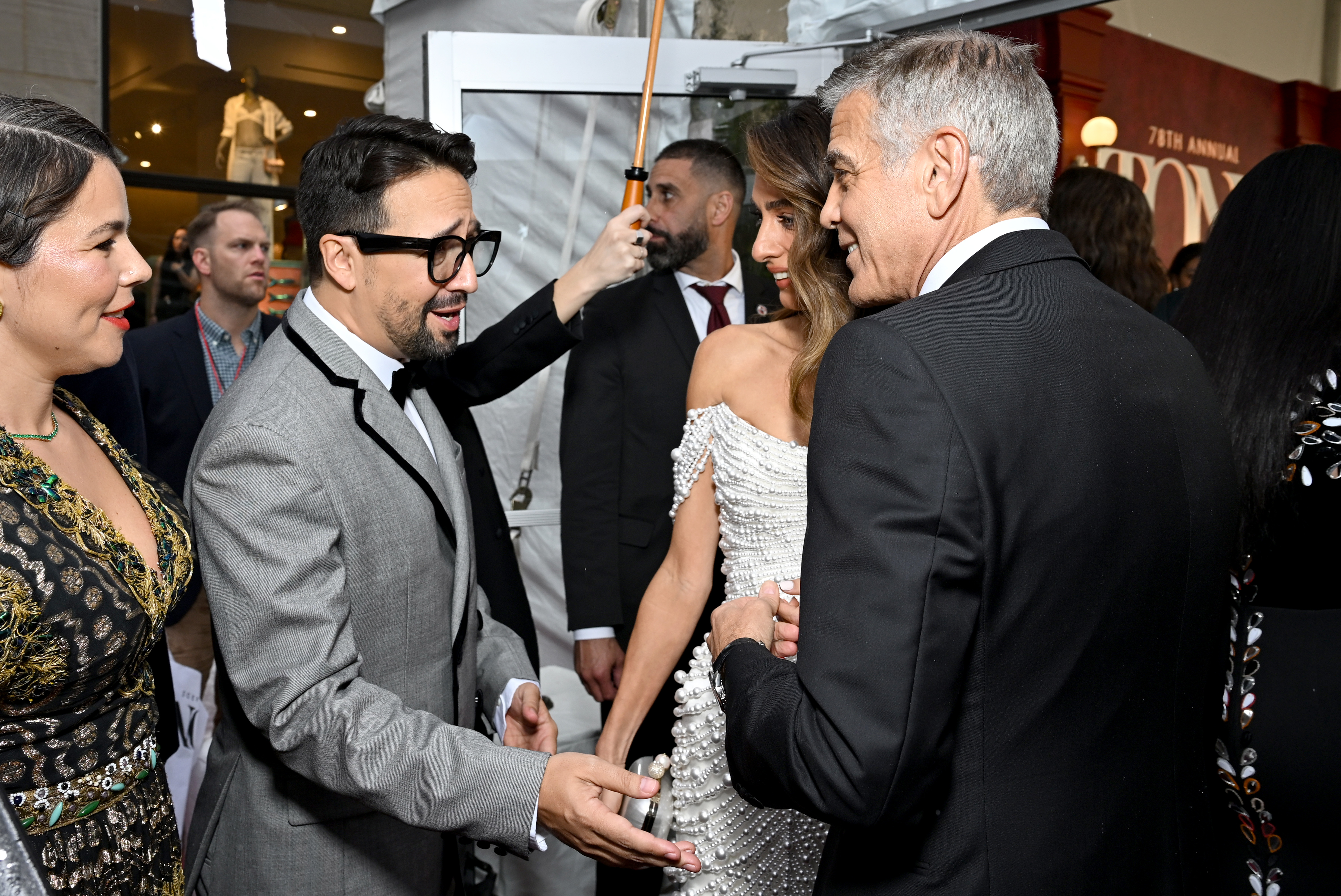 In a candid exchange, George and Amal Clooney engage in conversation with Lin-Manuel Miranda. Amal smiles warmly as George chats animatedly, creating a lively moment under the red canopy.