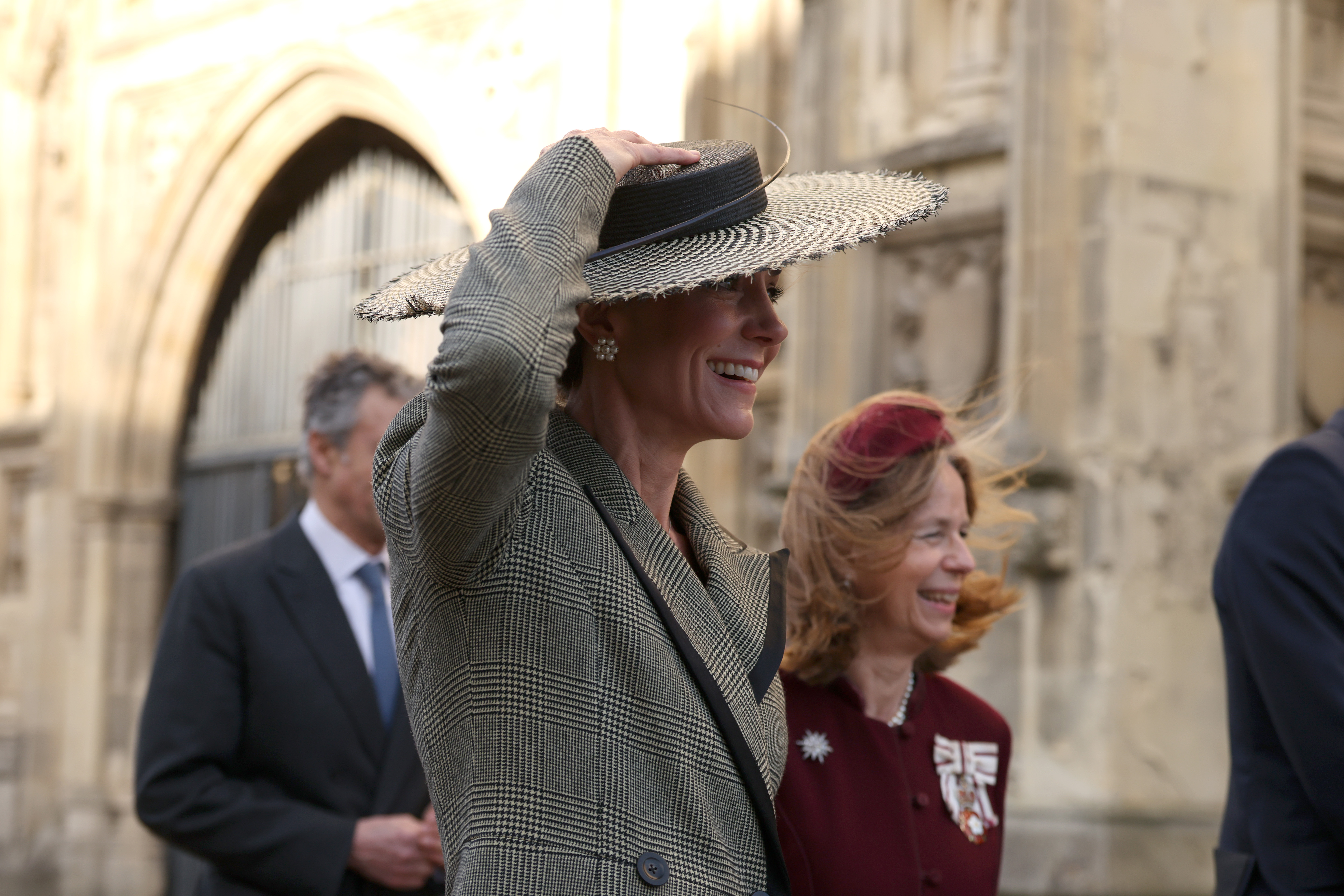 Both the Princess of Wales and her companion are caught mid-laugh as another gust sweeps through the cathedral grounds, sending hair flying in every direction. The Princess's pearl cluster earrings by Cassandra Goad — the same pair she wore to Prince Louis's christening in 2018 — are visible against her chignon, and the expression on her face confirms what the footage had already suggested — she found the whole thing just as funny as everyone else did.