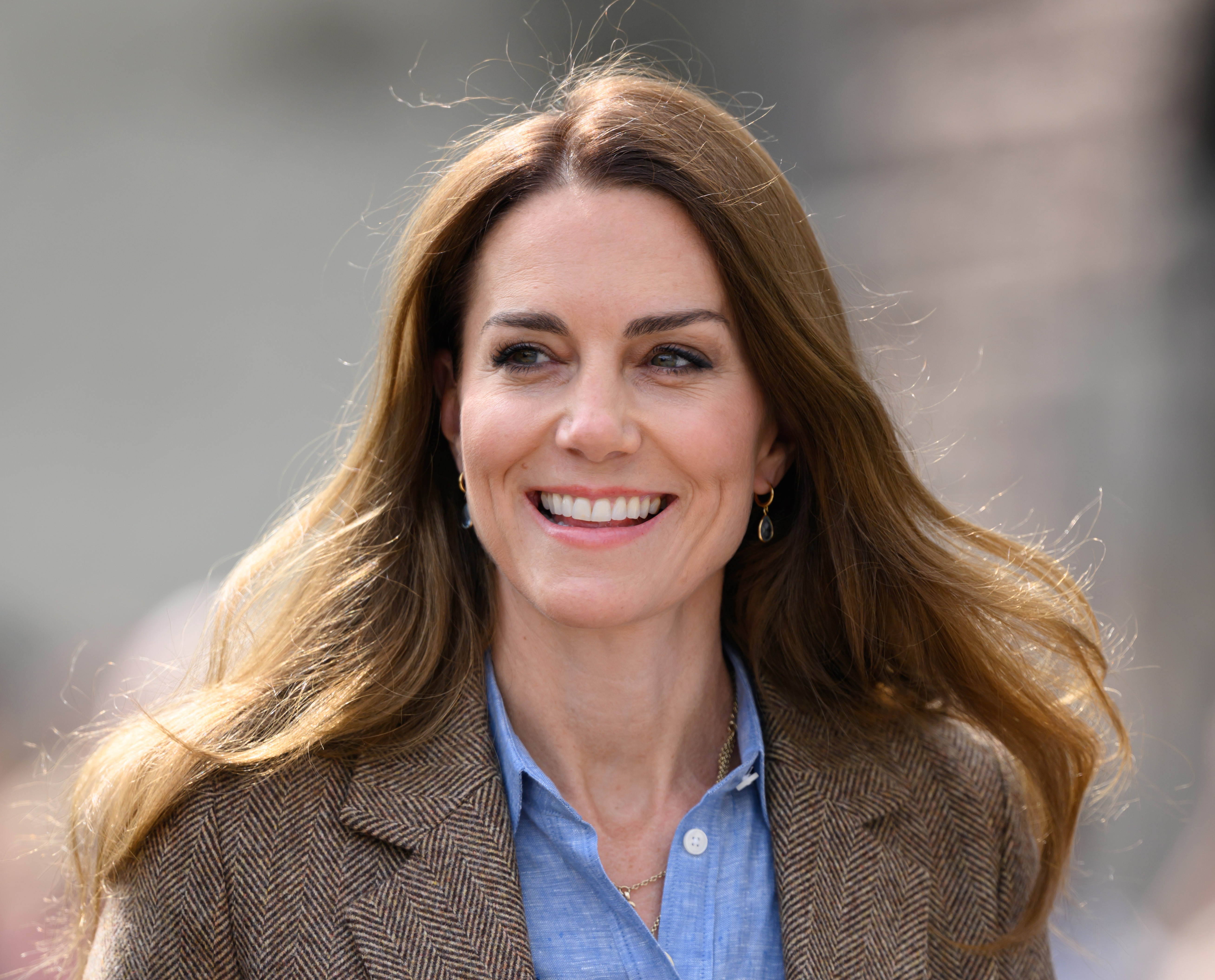 Catherine, Princess of Wales, looks radiant in a light blue button-up shirt layered under a brown herringbone blazer. Her long hair appears slightly lighter than usual, with warm golden highlights catching the sunlight, adding a subtle golden-yellow glow that softens her look