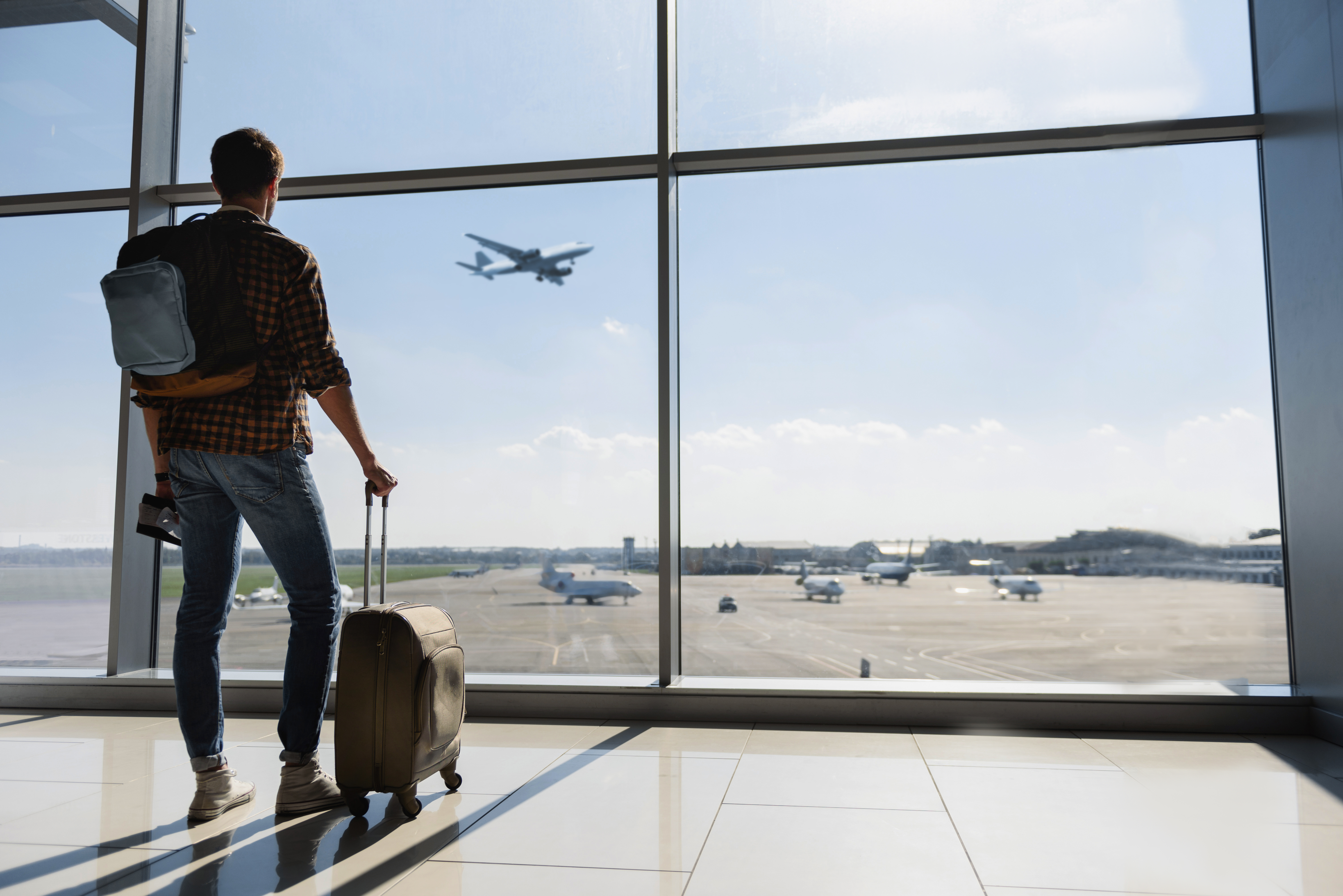 A young man with his luggage standing near an airport window and watching an airplane before departure | Source: Shutterstock