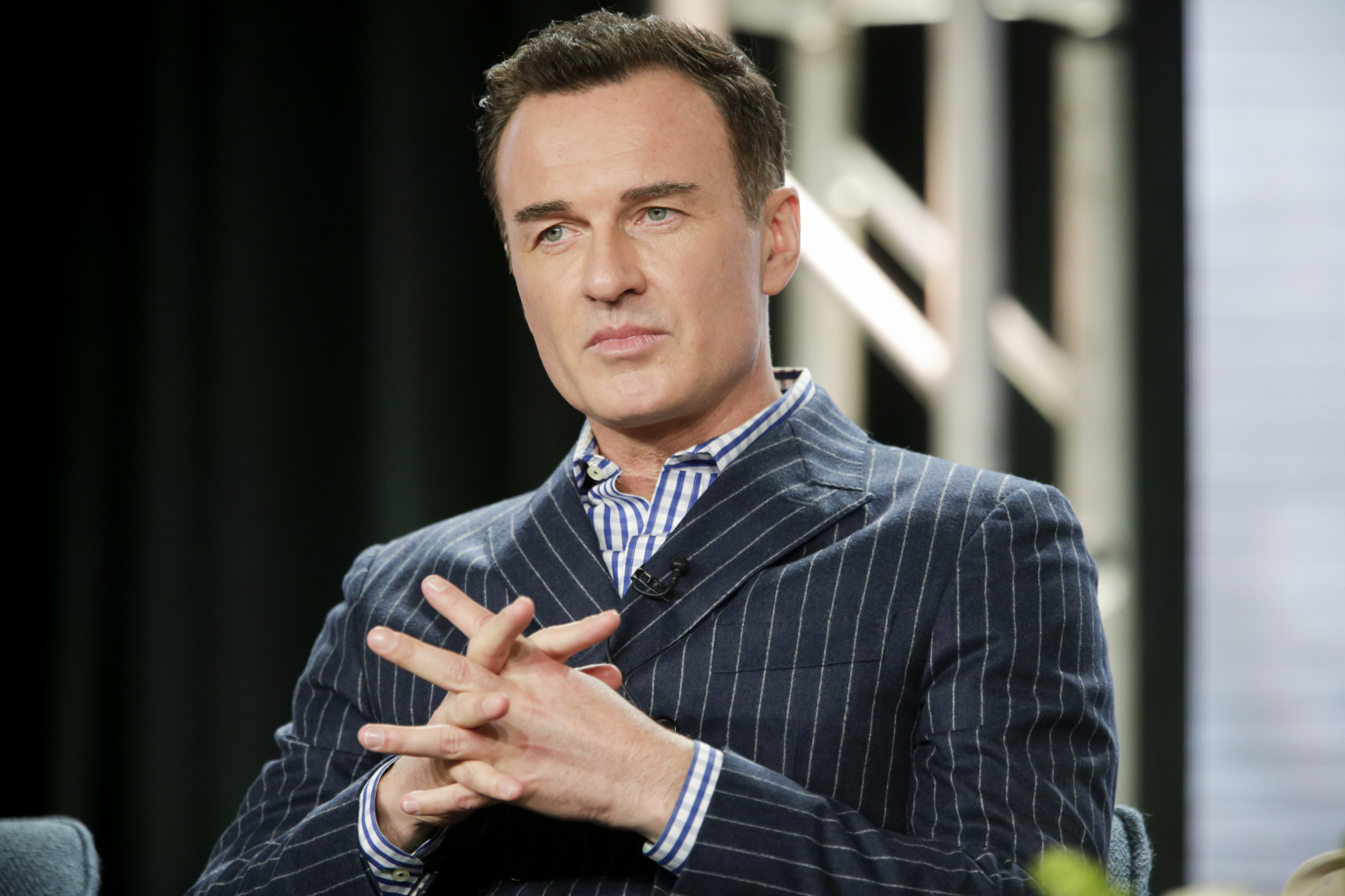 Julian McMahon is photographed during a panel discussion in 2020, wearing a navy pinstripe suit over a blue-and-white striped shirt. Seated with his hands interlocked, he maintains a composed, attentive expression. The soft stage lighting and industrial background suggest a formal media or television event.
