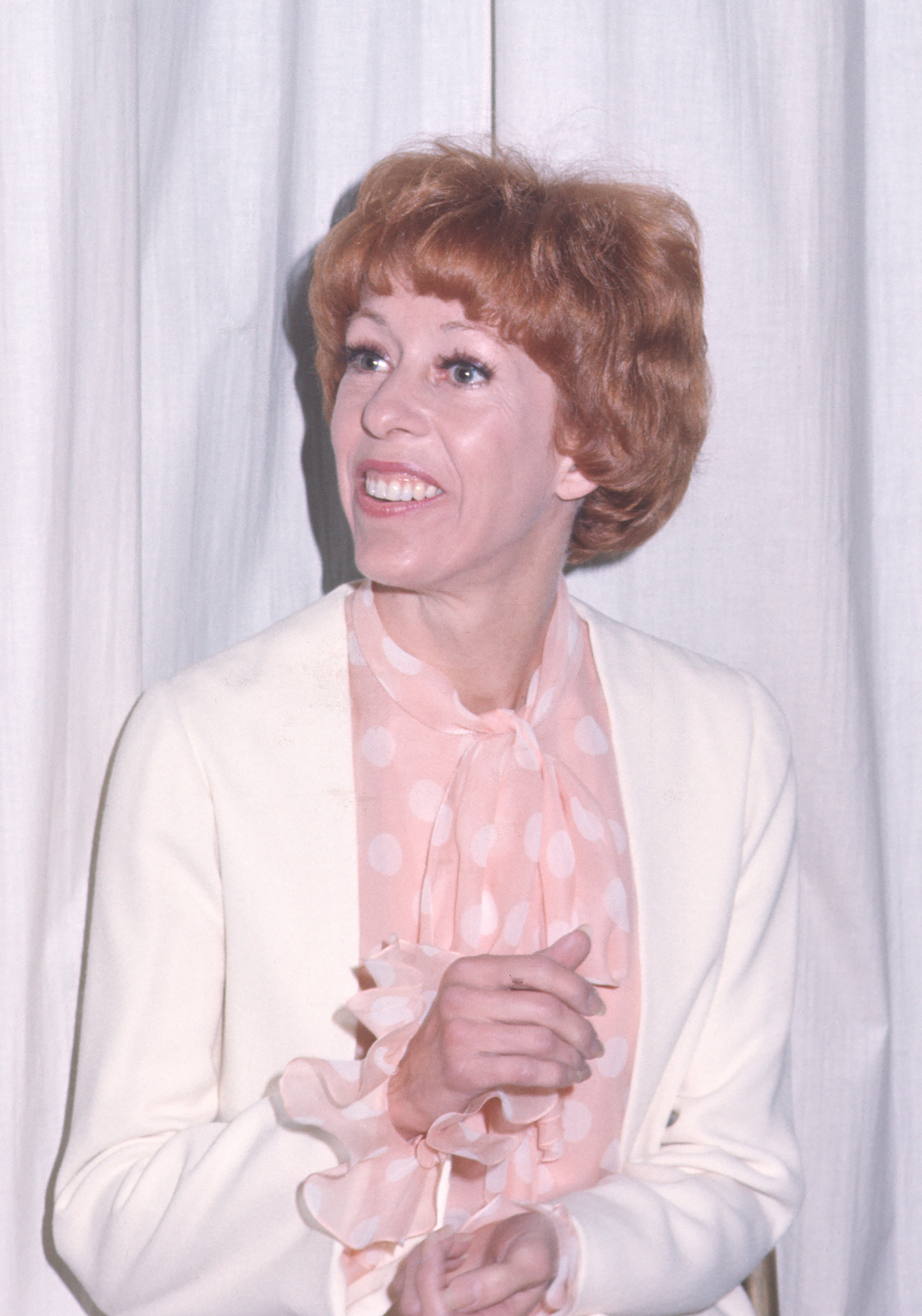 She wears a cream blazer over a soft pink blouse with a polka-dot pattern and a tied neckline. Her hands are loosely clasped as she turns slightly, looking off-camera.