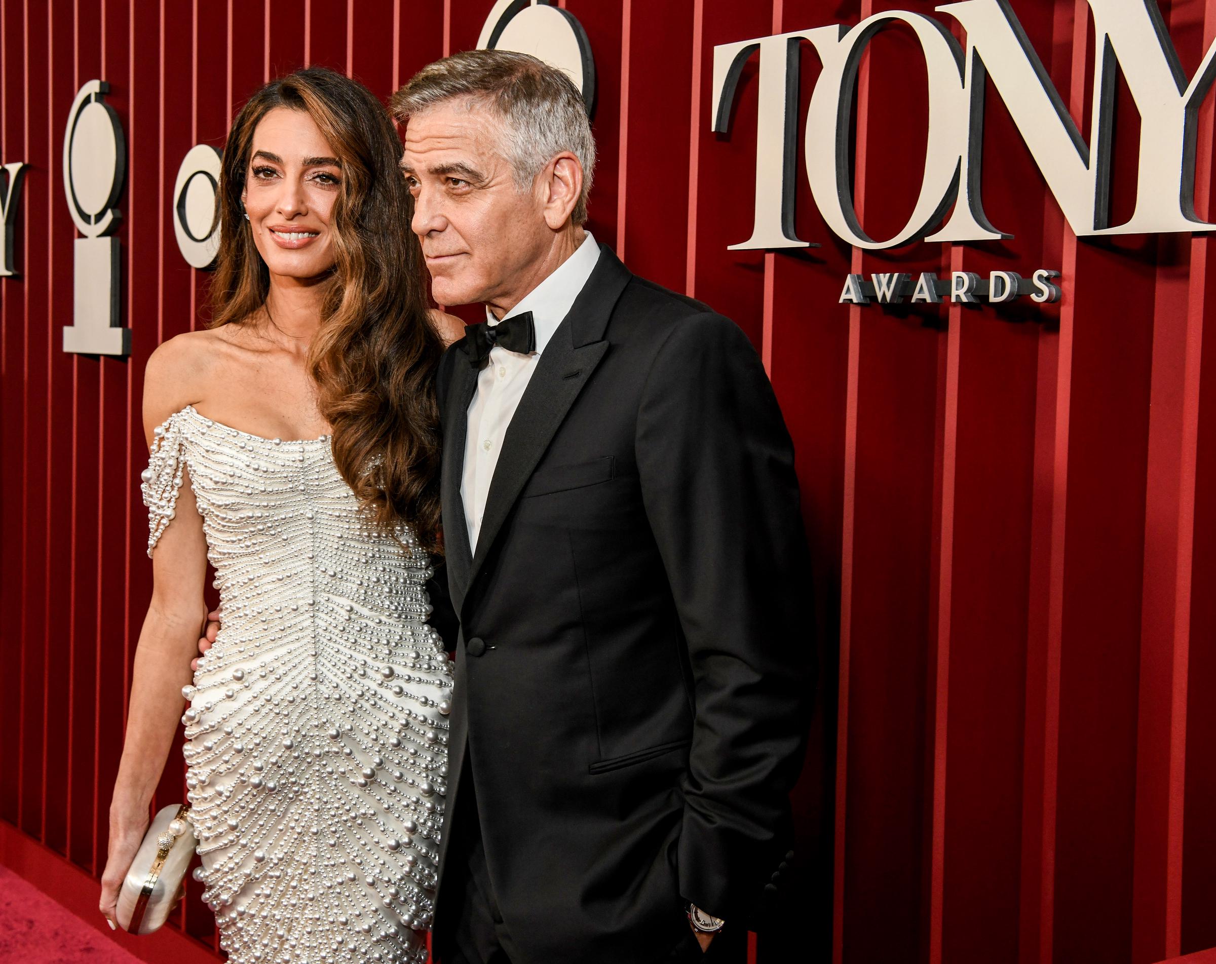 Amal Clooney stands with a soft smile, holding a pearl-studded clutch, while George Clooney stands beside her with his hands in his pockets.