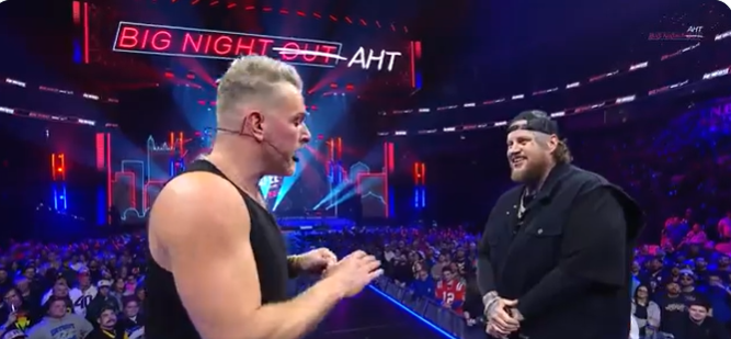 Jason "Jelly Roll"  is seen on stage with Pat MacAfee, smiling warmly under the bright arena lights. He wears a black jacket over a dark outfit, paired with a backward baseball cap, exuding a relaxed and approachable vibe. The energetic crowd and vibrant set design behind him highlight the lively atmosphere of the event.