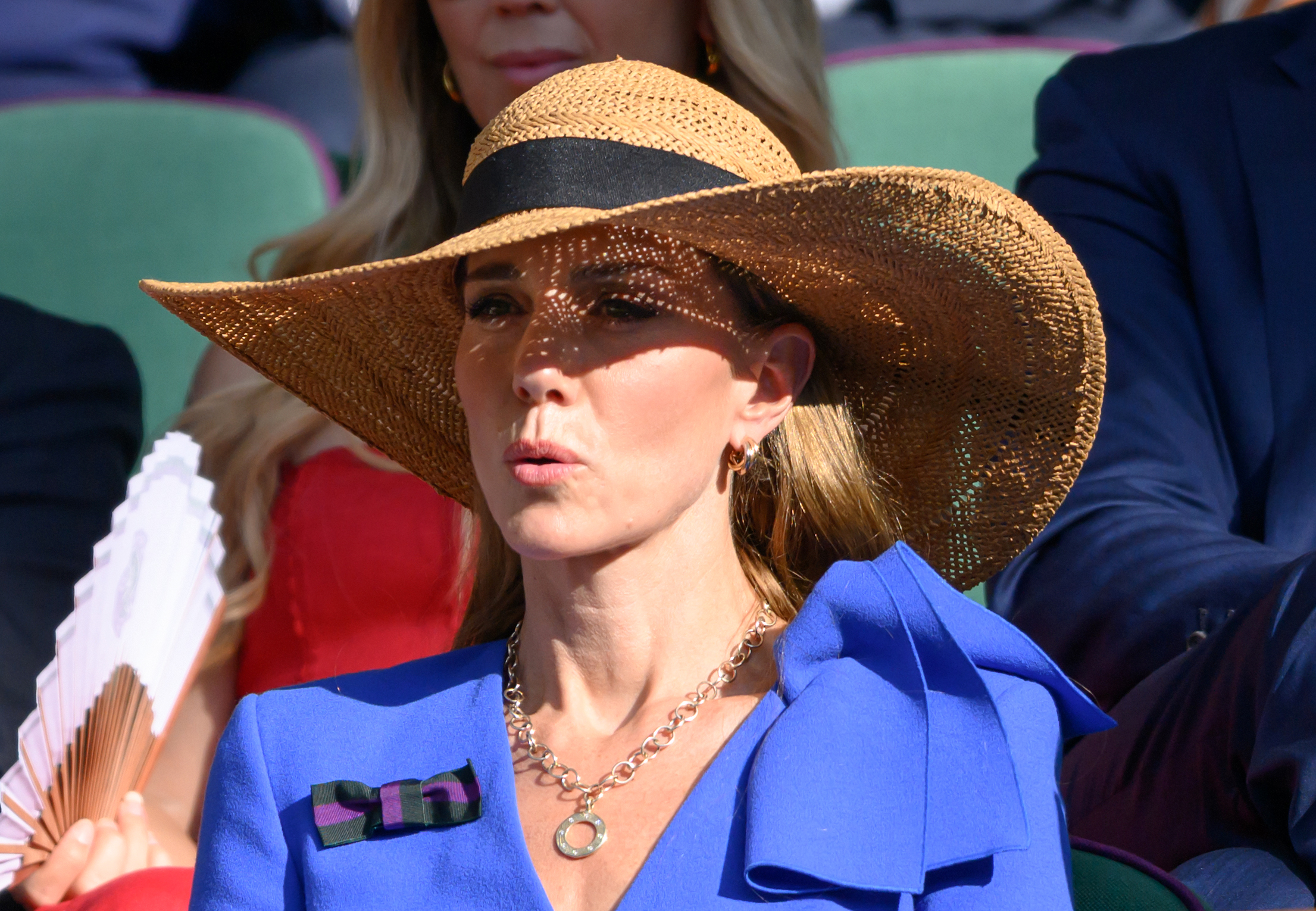 Princess Catherine reacts during a tense moment in the match while seated in the Royal Box. Her wide-brimmed hat shades her face as she fans herself against the summer heat.