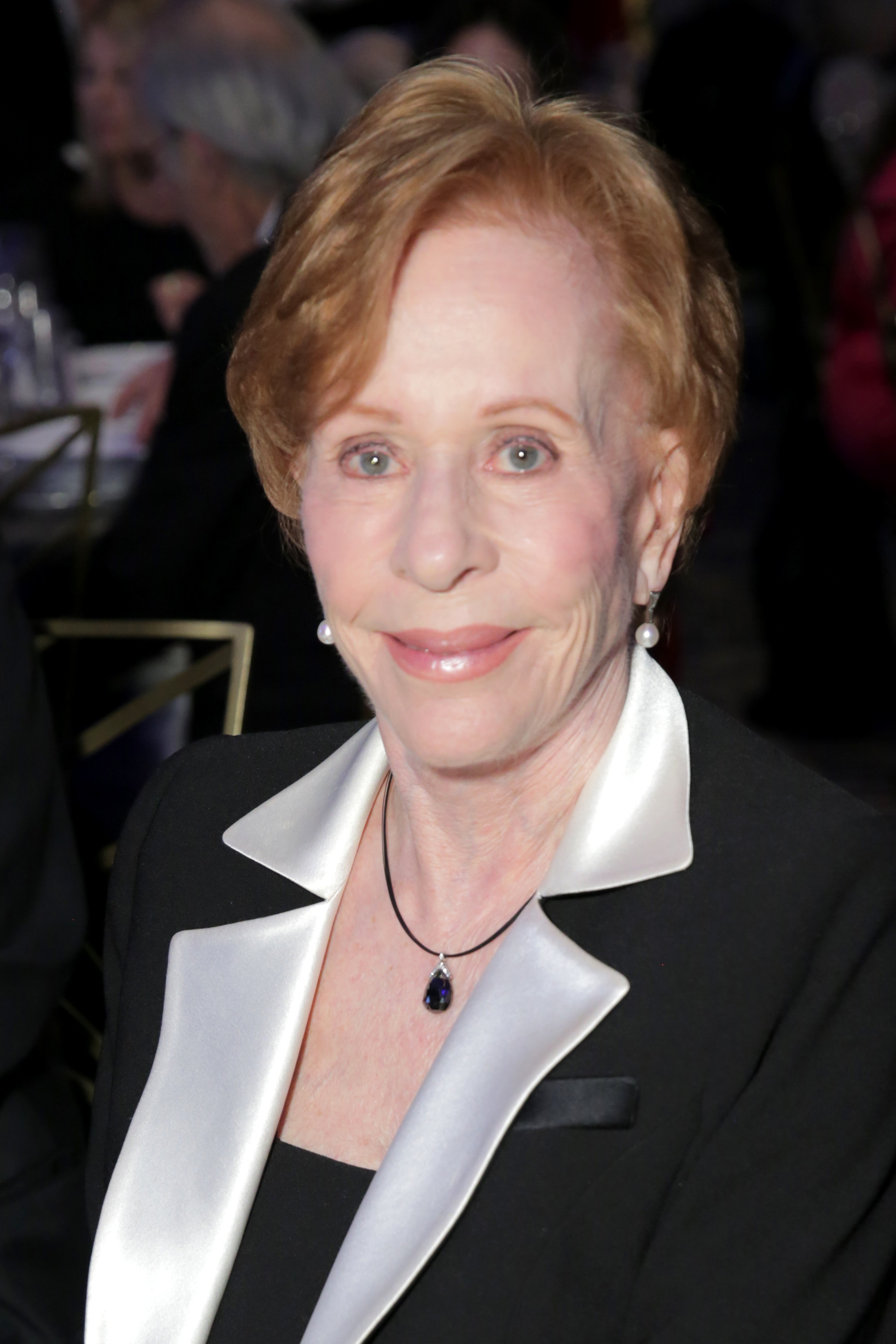 At a formal event, she wears a black outfit layered with a shimmering bronze-and-black sequined jacket. A small black handbag hangs from her shoulder, and she accessorizes with bracelets and a ring. She stands with hands gently clasped, while a table with bottles and another guest appear in the background.