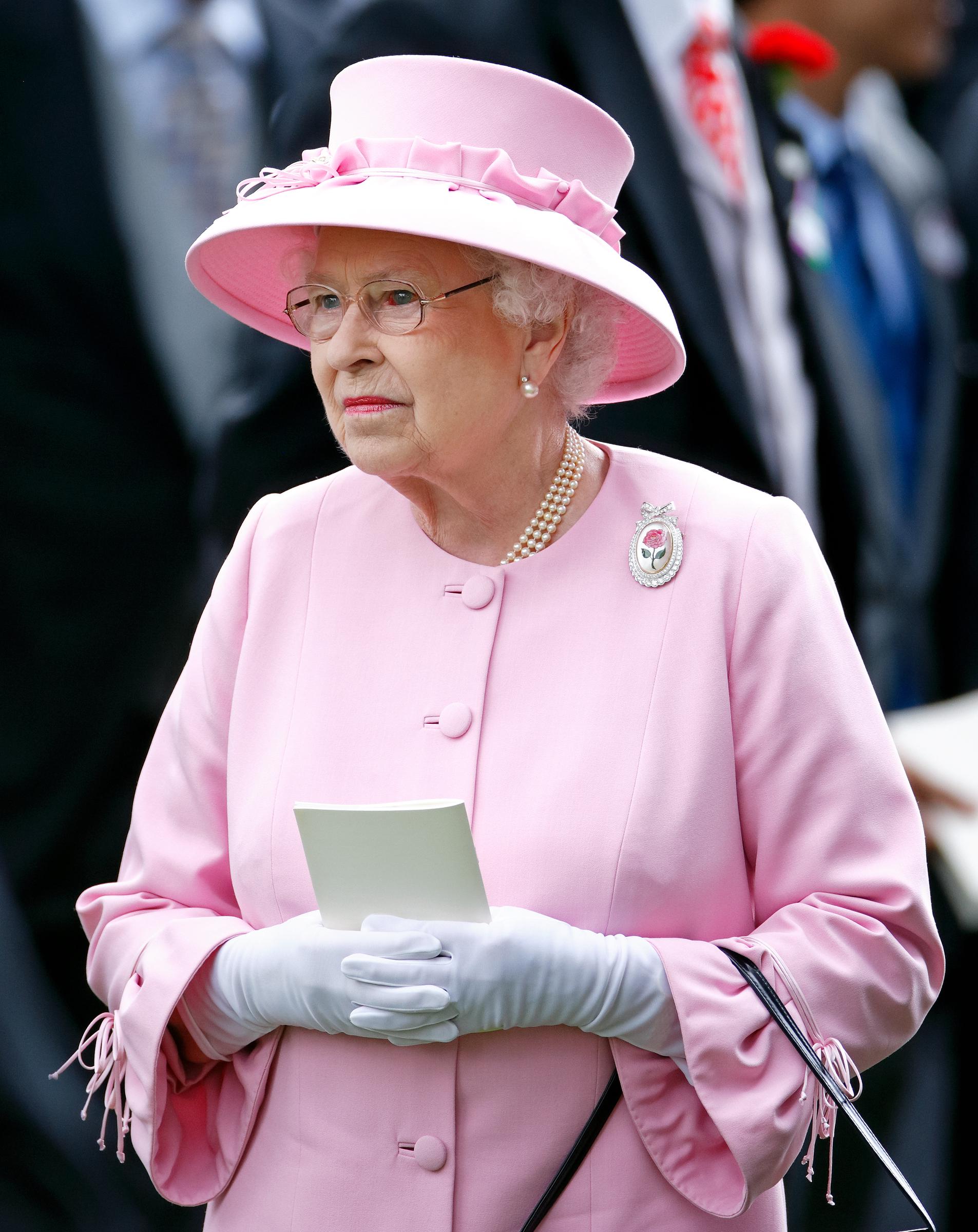 Holding an object and wearing soft white gloves, Queen Elizabeth II's elegance shines at the 2012 event. A visible red eye marks the moment, offering a rare glimpse of vulnerability amid her otherwise composed presence.