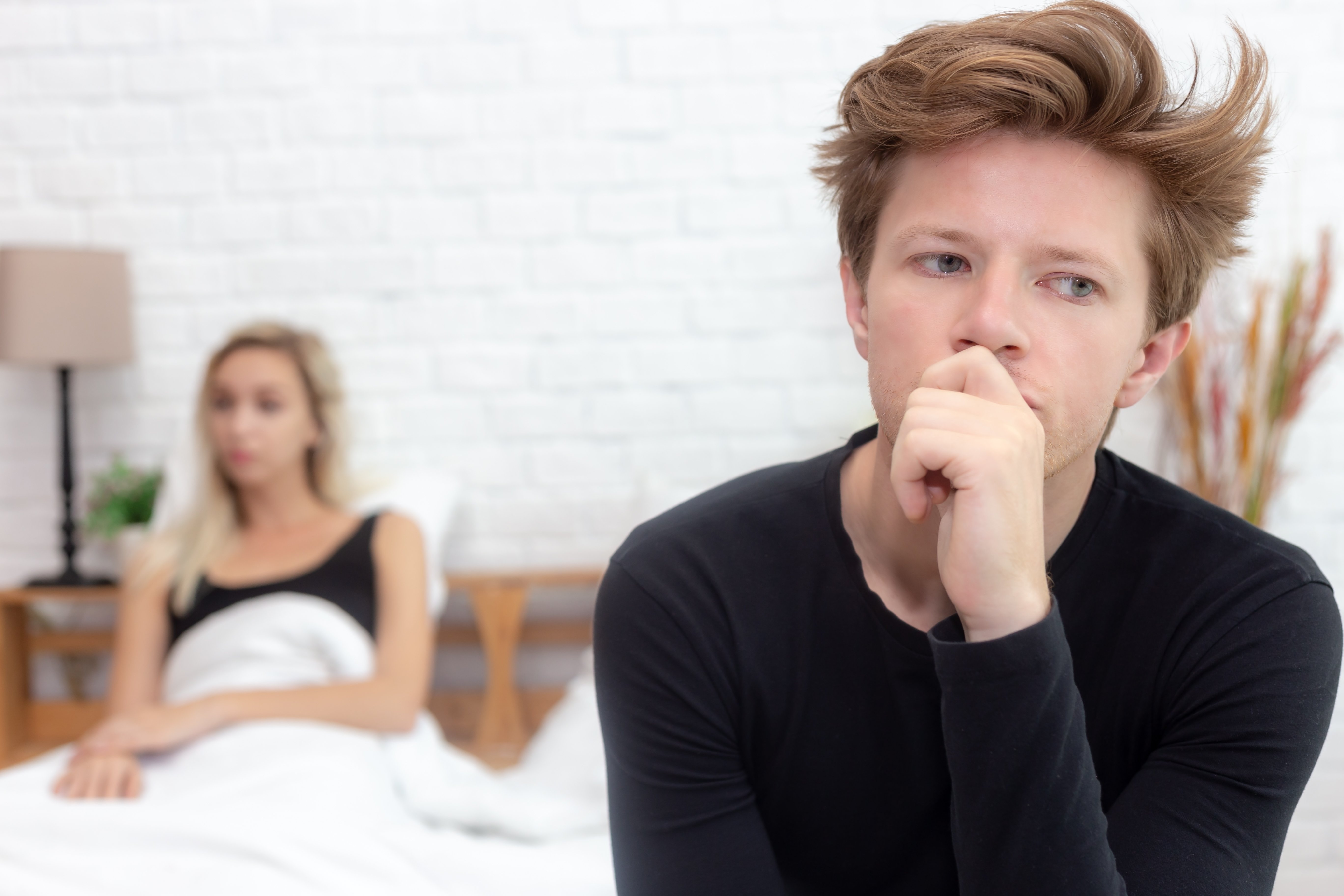 Hombre pensativo al pie de la cama con su pareja en el fondo || Fuente: Shutterstock Hombre pensativo al pie de la cama con su pareja en el fondo || Fuente: Shutterstock