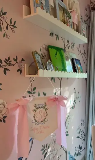 A delicate banner featuring the name Sofia Marie Scott hangs on nursery shelves filled with children’s books and keepsakes, adding a sentimental and personalized touch to the space. The scalloped edges and soft pink ribbons add extra charm. | Source: Instagram/alexmariedrum