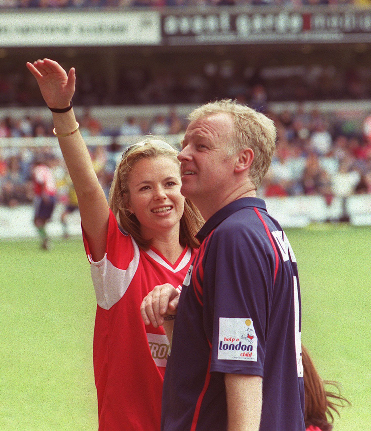 Holden waves to the crowd on the pitch at Stamford Bridge in London, wearing a red football shirt, as Dennis looks on beside her in a navy Help a London Child charity polo shirt. The pair attended the seventh annual Soccer Six celebrity tournament together.