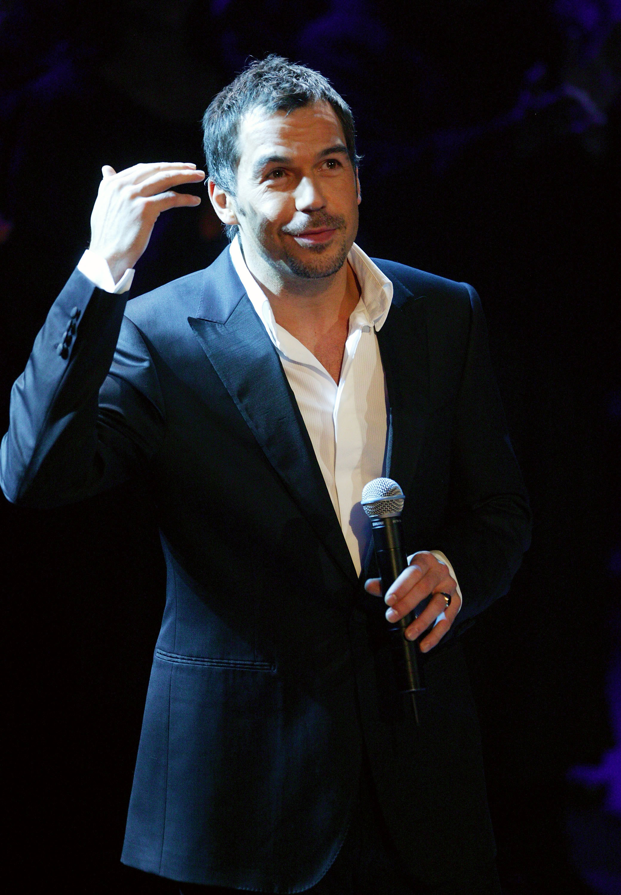 Brookstein works the crowd at the same Christmas Party, gesturing with one hand whilst clutching a microphone in the other, his dark hair slightly tousled and a knowing half-smile on his face.