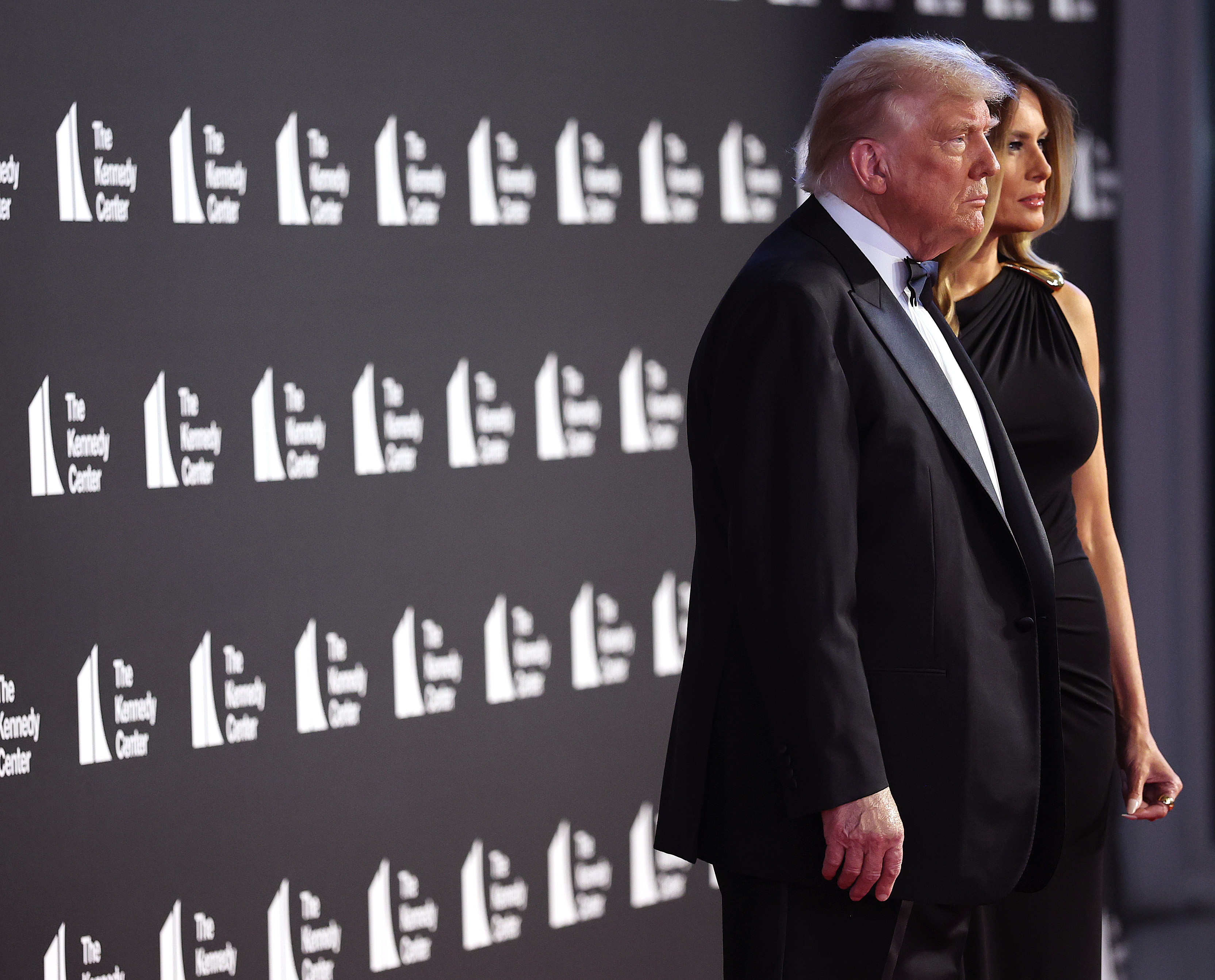 Donald and Melania stand side by side on the red carpet, facing forward with serious, composed expressions. The angle reveals the sleek silhouette of Melania’s gown, its fabric smoothly fitted through the waist and hips.